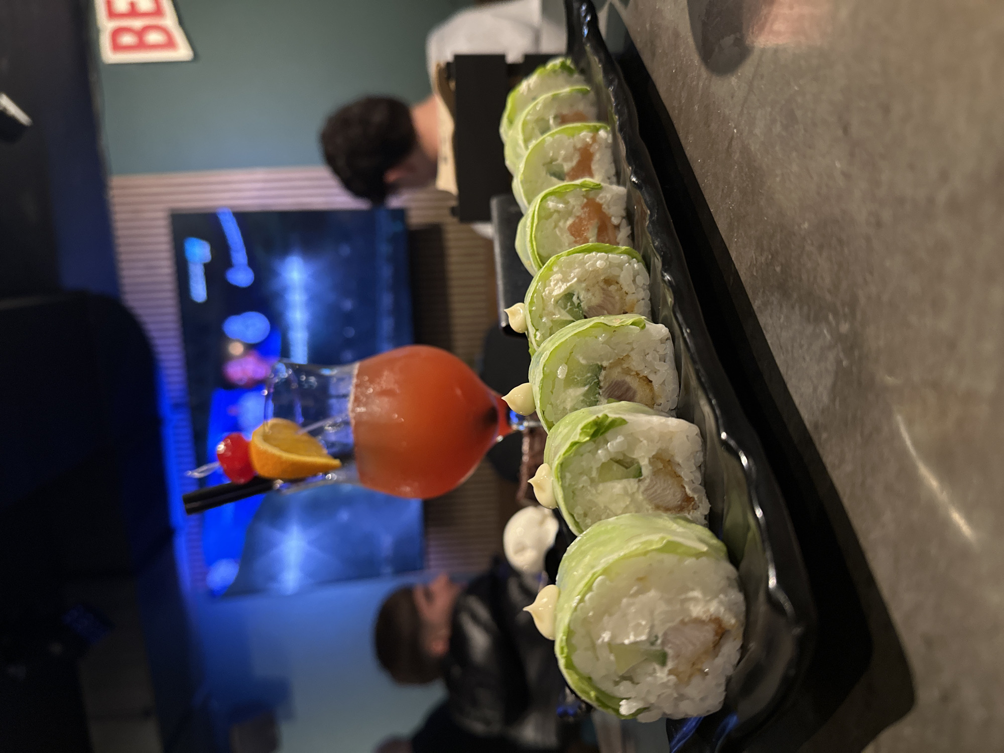 The image shows a close-up of a platter of sushi rolls and a cocktail on a reflective surface, seemingly a table. The sushi rolls are wrapped in lettuce instead of seaweed and topped with a small dollop of mayonnaise. The cocktail is a reddish-orange color and garnished with a cherry, an orange slice, and a black straw. In the background, reflections of people are visible along with a television screen and a sign with the letters "BE" in red. The location is possibly a restaurant in Burgas, Bulgaria. The people in the background appear to be dining or working in the restaurant.