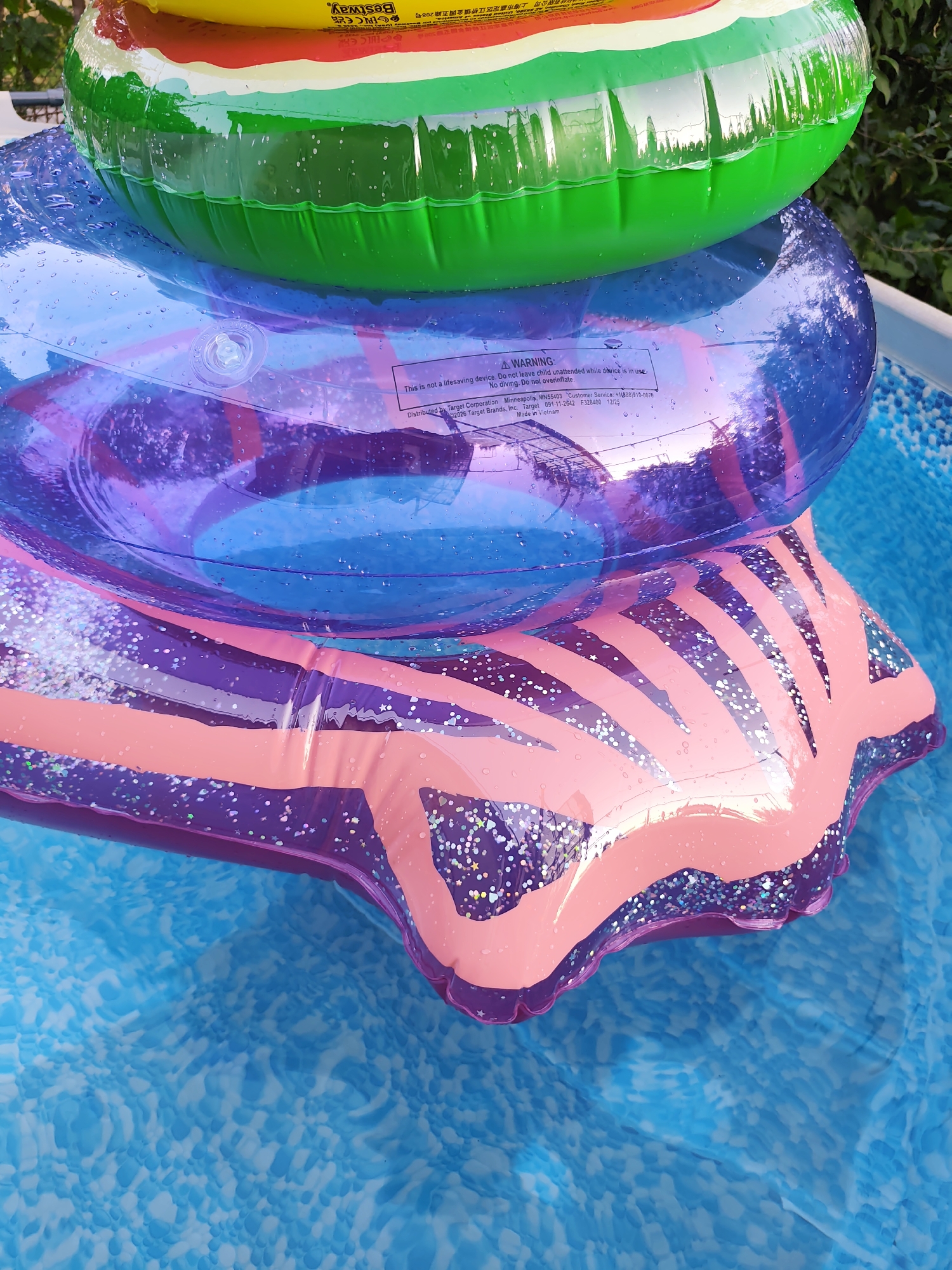 The image shows a stack of inflatable pool toys floating in clear blue water. The top toy is a green, yellow, and red ring, resembling a watermelon slice. Beneath it is a transparent purple ring with glitter. The bottom toy is a pink and purple star-shaped inflatable with glitter. The water has a patterned texture. The background is slightly out of focus, showing green foliage and possibly a fence. The scene appears to be set in an outdoor swimming pool. There is a warning label visible on the purple inflatable. The image does not contain any specific geographical markers that would indicate its location in Durlești, Moldova.