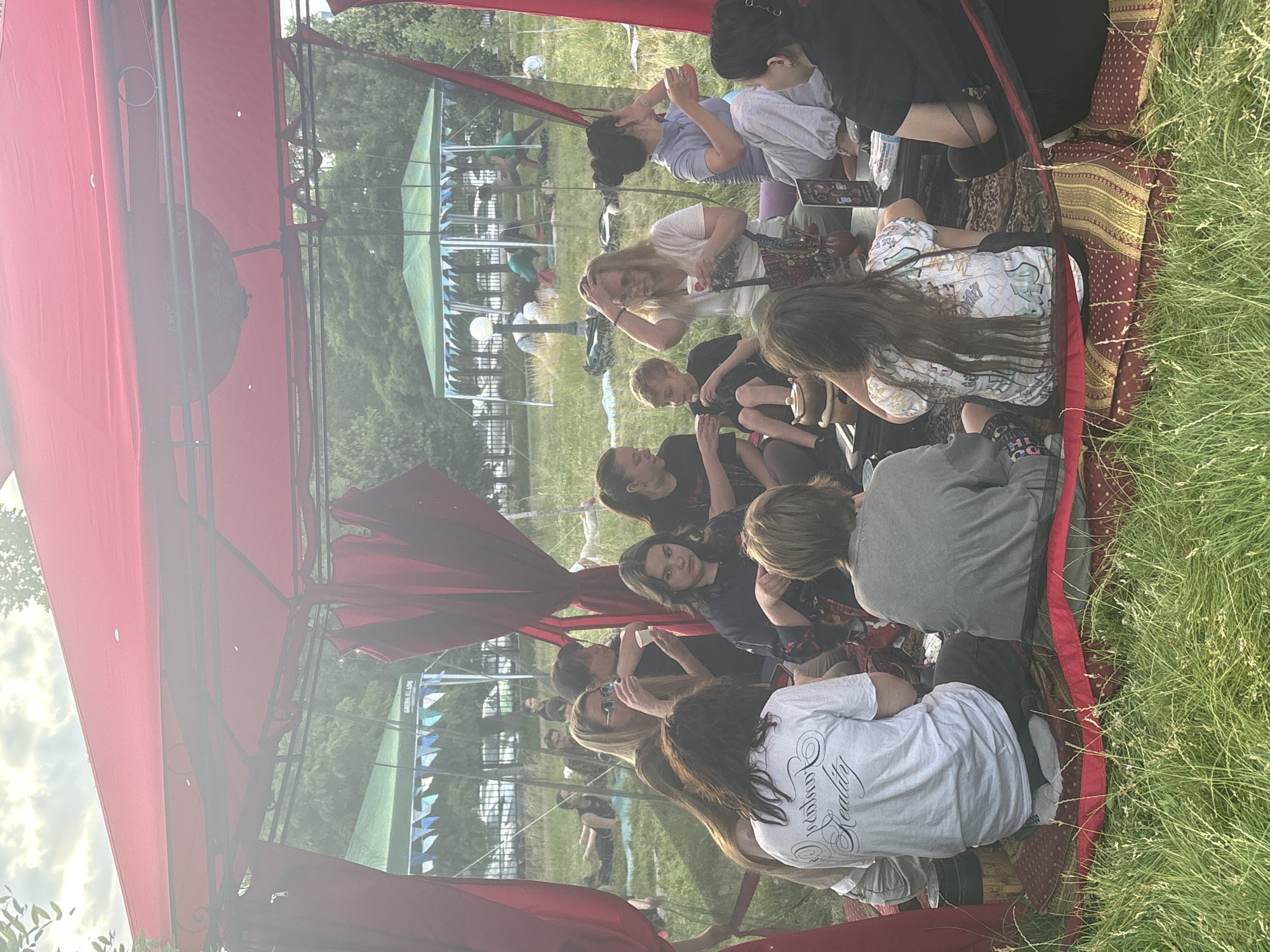 An outdoor gathering is taking place under a red canopy tent. A group of approximately ten individuals, including adults and one child, are seated on cushions and rugs on the grass, some holding cups. The setting appears to be a park or open field, with trees and other tents visible in the background. The overall atmosphere is relaxed and social. The image does not provide enough information to determine its location within the city of Astrakhan, Russia.