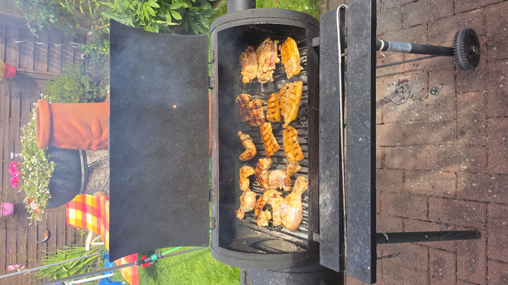 In a backyard setting, likely in Hamm, Germany, a black barbecue grill is open and filled with various cuts of raw and partially cooked chicken. The chicken pieces are arranged on the grill grates, with some showing browning and grill marks, indicating they are in the process of being cooked. Smoke is visible rising from the grill, suggesting active cooking. The grill is situated on a brick patio. In the background, there is a wooden fence, green grass, and foliage. A lounge chair with a colorful striped cushion is visible to the left of the grill, along with several potted plants and a terracotta planter with a face design. The scene depicts a typical outdoor grilling event.