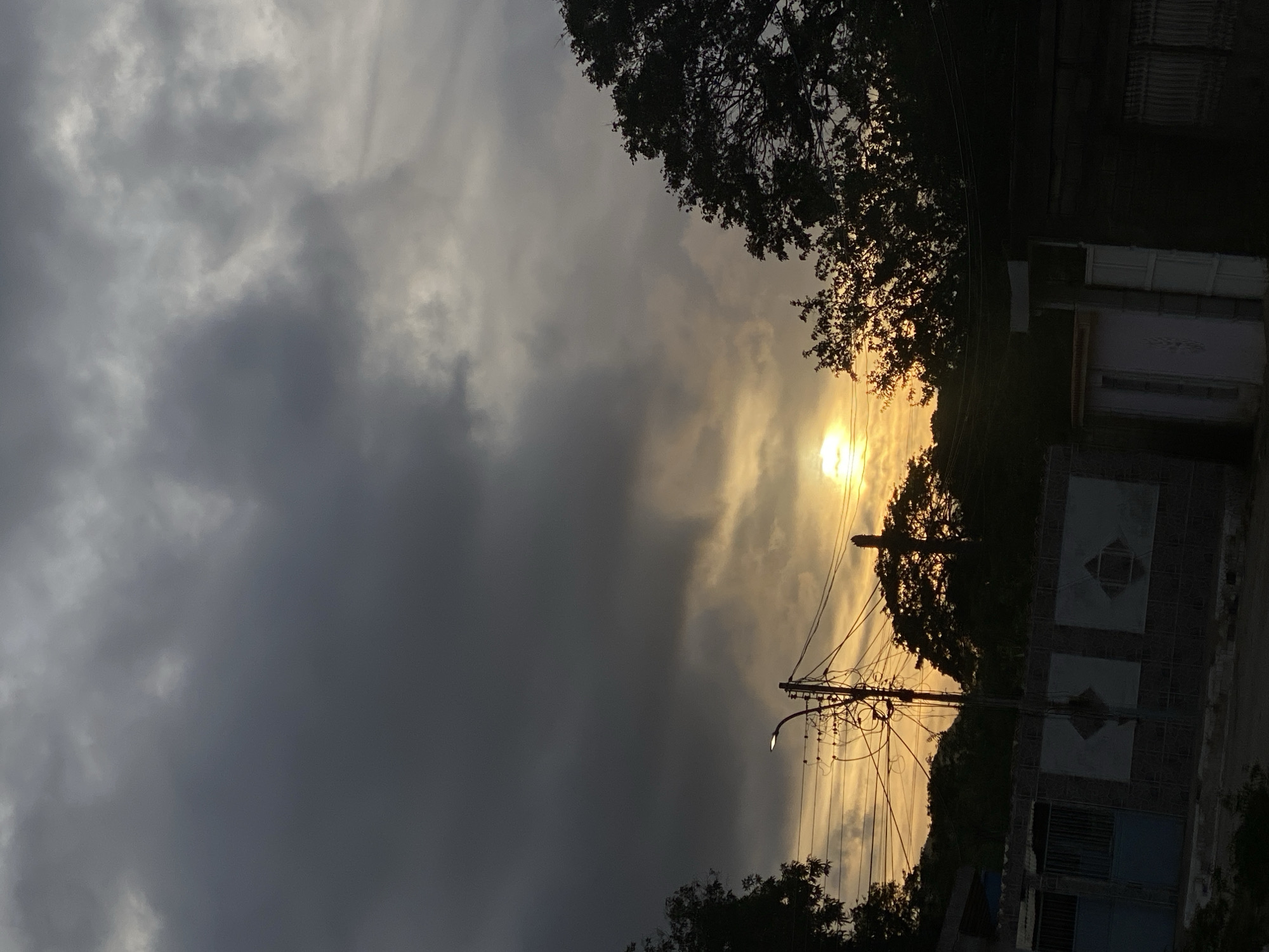 The image depicts an outdoor street scene in Juan Griego, Venezuela, at either sunrise or sunset. The sky, occupying the upper two-thirds of the frame, features dense, dark gray clouds in the upper left, transitioning to lighter gray and warm orange-yellow hues near the horizon. A prominent, bright yellow-orange light source, consistent with a low sun, is visible near the center-right horizon, partially obscured by dark, silhouetted tree foliage.
In the midground, a dark utility pole, equipped with a street light fixture pointing slightly upwards, is situated to the left of center. Numerous intertwined electrical and communication wires extend horizontally from this pole across the frame. Additional silhouetted trees and dense foliage are visible behind the utility pole and extensively on the right side.
The foreground reveals elements of residential properties. On the left, a dark-colored wall or fence is present, featuring three lighter rectangular panels, each containing a dark diamond-shaped insert. A partially visible blue metal gate is located behind this wall section. On the right side of the foreground, dark building facades are discernible, along with a light-colored door or gate and a dark fence composed of vertical slats. The immediate ground level is largely obscured by shadow. No individuals or specific activities are visible within the frame.