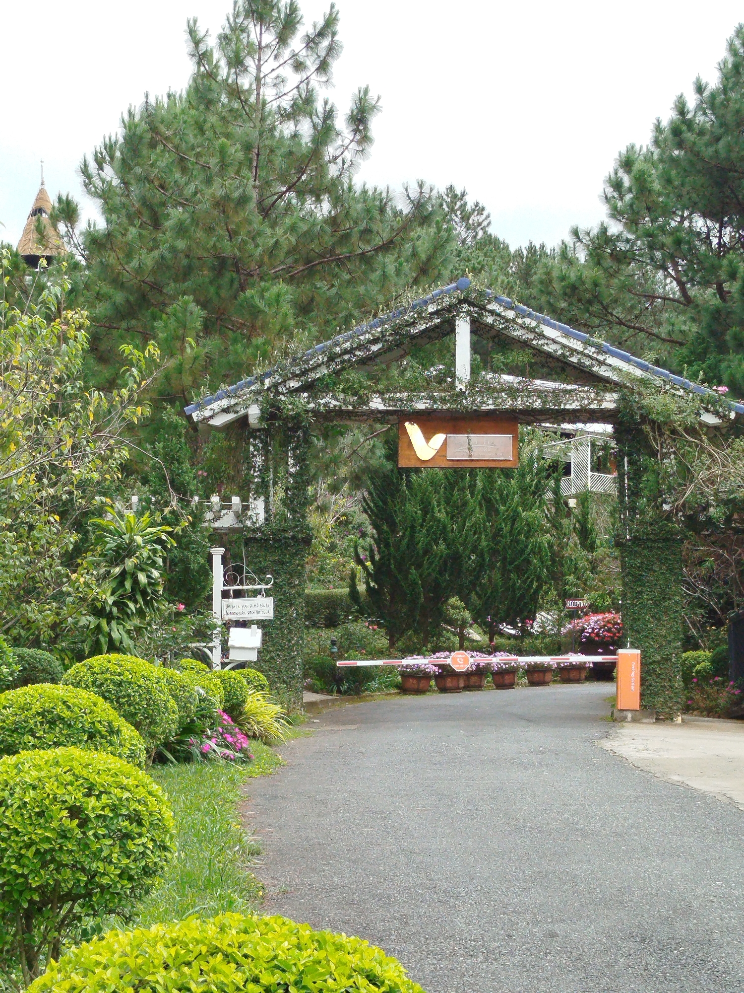 The image depicts the entrance to a property in Lâm Viên - Đà Lạt, Vietnam. A white, arched gateway with a blue-tiled roof, covered in ivy, marks the entrance to a paved road. A wooden sign with a yellow "V" logo and text hangs above the gateway. A barrier gate is lowered across the road, with several potted plants lining the sides. To the left of the gateway, a white signpost stands with a decorative white sign. Lush green trees and manicured bushes surround the entrance, with a building visible in the background through the gateway. The overall impression is of a well-maintained, scenic entrance to a resort or estate.