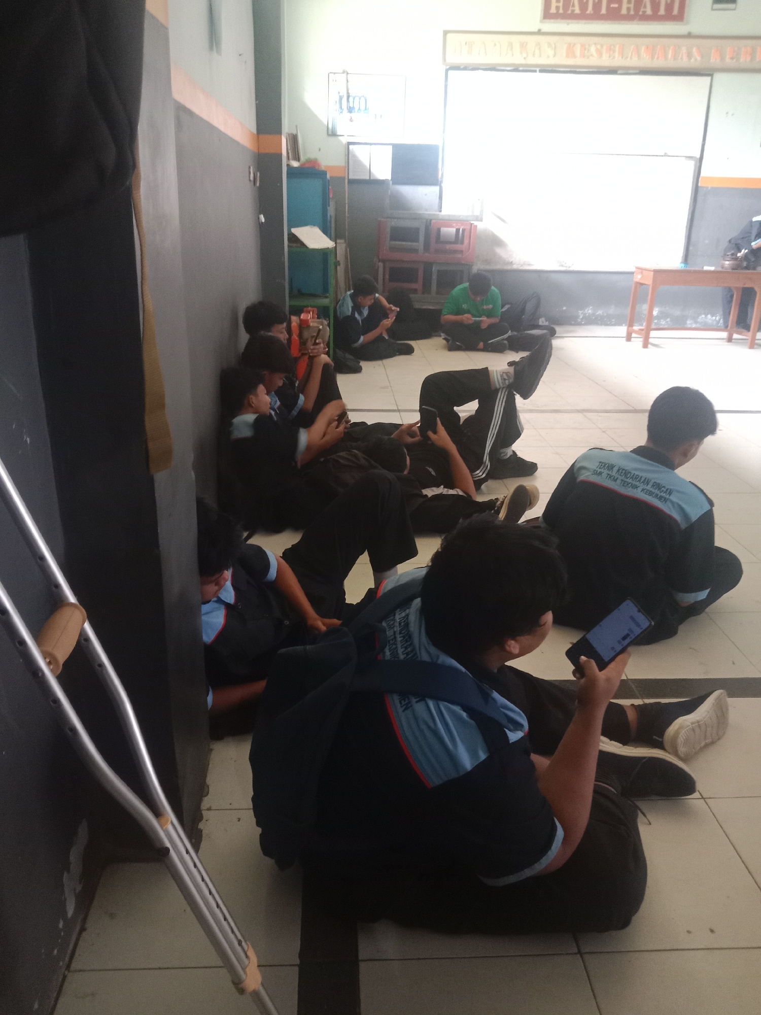 The image depicts a group of young males, presumably students, sitting on the floor in an indoor environment. Several individuals are holding and looking at their mobile phones, suggesting they are engaged with their devices. One person is positioned near a pair of crutches, indicating a possible injury or mobility issue. The background reveals what appears to be a classroom or common area with a whiteboard, wooden table, and storage cabinets. The writing on the whiteboard and the uniforms worn by some individuals suggest an educational institution, possibly SMK TKM Kebumen, located in Kebumen, Indonesia. The overall scene conveys a relaxed, informal atmosphere, with the students seemingly waiting or taking a break.