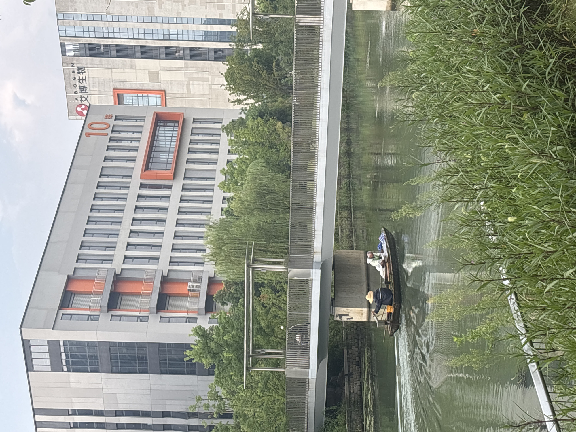 The image depicts a serene canal scene in the Suzhou Industrial Park, China. In the foreground, lush green reeds and vegetation line the canal bank, with a modern pedestrian bridge spanning across the water. The bridge features a silver metal railing and a covered area with seating, suggesting it's a place for people to rest and enjoy the view. Underneath the bridge, a small boat with two individuals is navigating the canal. One person, wearing a straw hat, appears to be steering the boat, while the other is seated behind. The boat is leaving a wake, indicating movement. In the background, imposing modern buildings rise, one of which prominently displays the number "10" and an emblem with Chinese characters and "ABOGEN." The architecture of the buildings is characterized by large windows, grey facades, and striking orange accents, hinting at a blend of industrial and corporate design within the park. The water of the canal is calm and reflects the sky and surrounding structures. The overall atmosphere is one of a tranquil, developed urban environment.