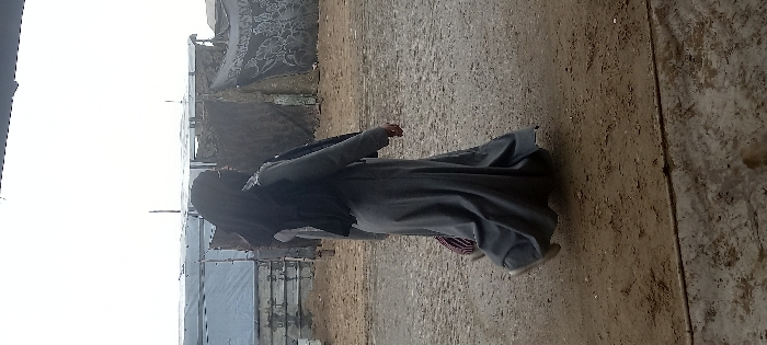The image depicts a solitary individual walking from left to right across a barren, unpaved area in Khan Yunis, Palestinian Territory. The person is dressed in a long, grey full-length garment, a black jacket or vest, and a black hijab covering the head, with light-colored footwear visible on the left foot. A striped bag, featuring red, white, and dark colors, is carried over the left shoulder, partially obscured. The ground consists of uneven dirt and gravel, with a dark, horizontal linear object traversing the lower foreground. In the background, a series of temporary structures are visible. To the left, structures appear to be constructed from light-colored panels or sheeting. To the right, structures are formed from dark fabric or tarpaulins, some displaying white patterns, supported by vertical poles. The sky above is brightly lit or overcast, providing diffuse illumination to the scene. No other individuals are present or interacting in the frame.