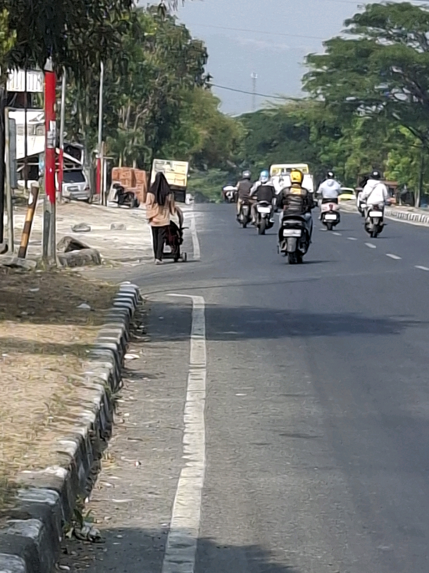 The image depicts a street scene in Sukabumi, Indonesia. A woman in a peach-colored top and black headscarf is walking on the sidewalk, pushing a stroller. In the background, several people are riding motorcycles on the road. The road is paved and has white lane markings. Trees line the sides of the road, and there are some buildings and structures visible on the left side of the image. The sky is clear and bright. The overall scene suggests a typical day in a bustling Indonesian city.