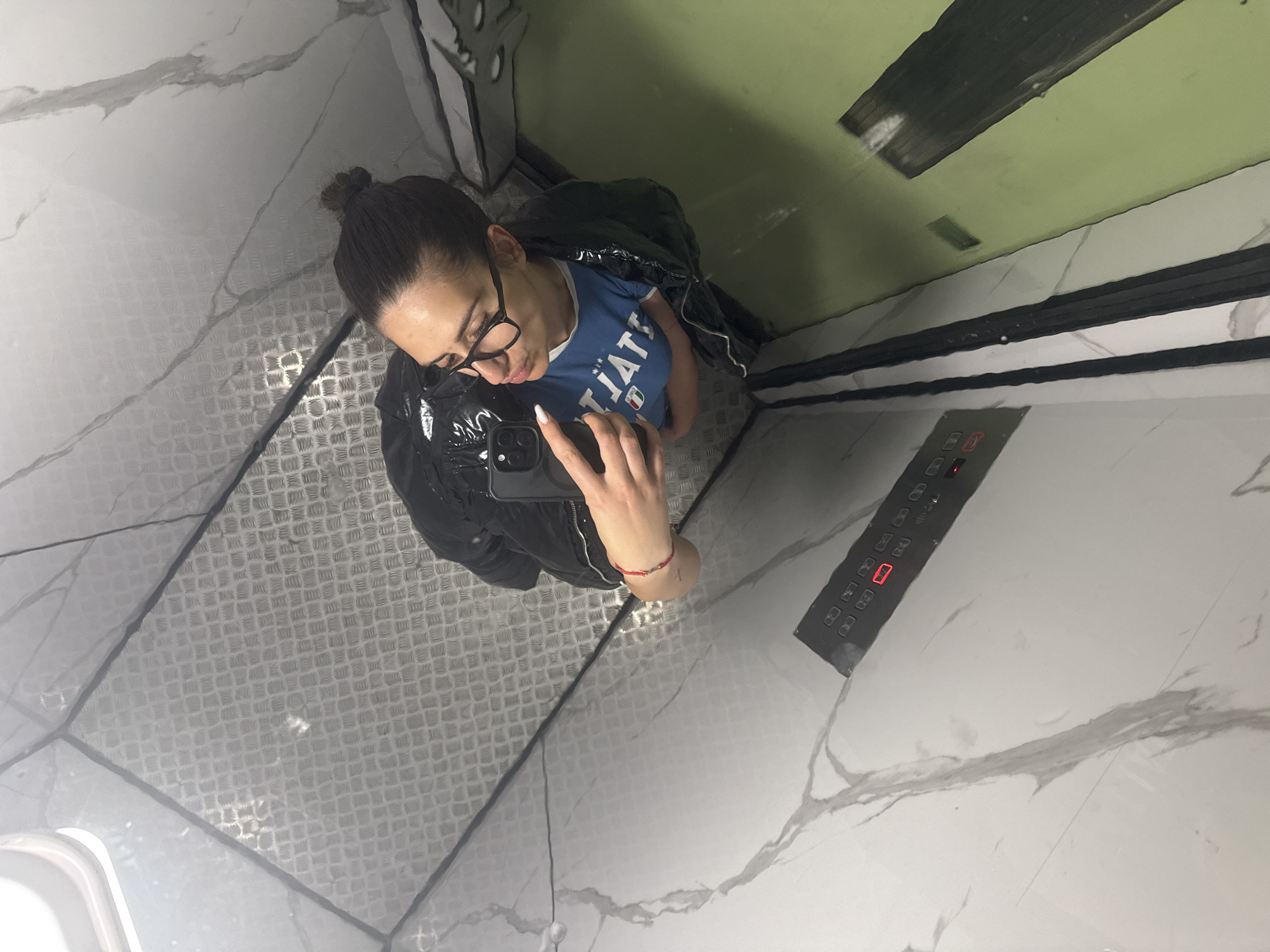 A woman wearing glasses, a blue t-shirt, and a black jacket takes a selfie with a black smartphone in an elevator. The elevator interior has gray patterned floors and white marble-patterned walls. A control panel with illuminated red numbers is visible on the right wall. The lighting is bright. Sofia, Bulgaria is not discernible from the image.