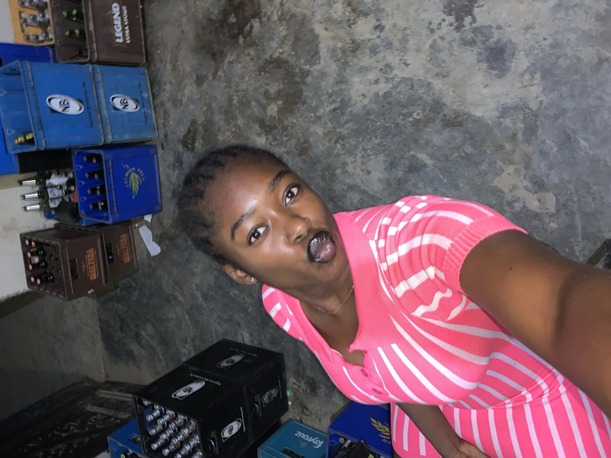 A woman in a pink and white striped dress is taking a selfie. She has dark hair pulled back from her face, dark eyes, and dark lipstick, and she is sticking out her tongue. She wears a gold necklace. The background shows stacks of crates, some containing bottles. Some crates are marked with "Legend Extra Stout," "Nigerian Breweries Plc," and "Fayrouz." The floor appears to be concrete. The location is implied by the presence of Nigerian products and a reference to the "New Stadium, Nigeria" in the prompt.