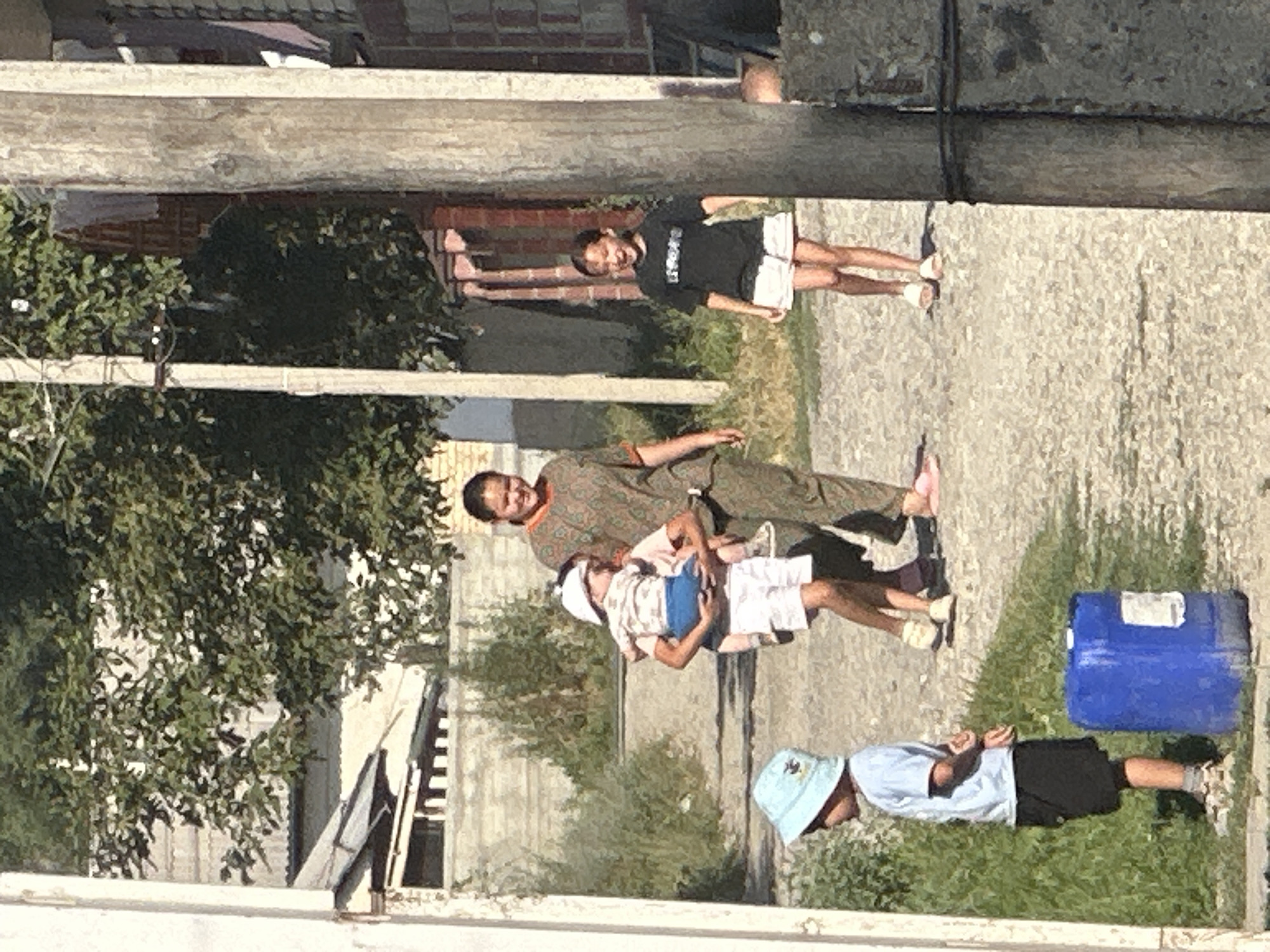A medium shot captures a sunny, outdoor scene on a dirt path in a residential area of International, Kyrgyzstan. In the foreground, a young boy wearing a light blue t-shirt, black shorts, and a light blue bucket hat stands facing left. To his right and slightly behind him is a large blue plastic barrel.
Moving further back and to the center, a woman in a patterned green and brown tunic is walking, carrying a child on her hip. The child is wearing a white hat, a white and patterned shirt, and white shorts. Behind them, a younger girl in a black t-shirt with white lettering and white shorts walks towards the viewer, smiling and with her mouth open.
The background consists of buildings with some foliage and trees. A light-colored wall with a fence is visible behind the children in the center. A weathered wooden utility pole stands on the right side of the frame, with wires running between it and another pole to its left. The ground is a mix of dirt, gravel, and patchy green grass. The lighting suggests it is daytime, with bright sunlight casting shadows.