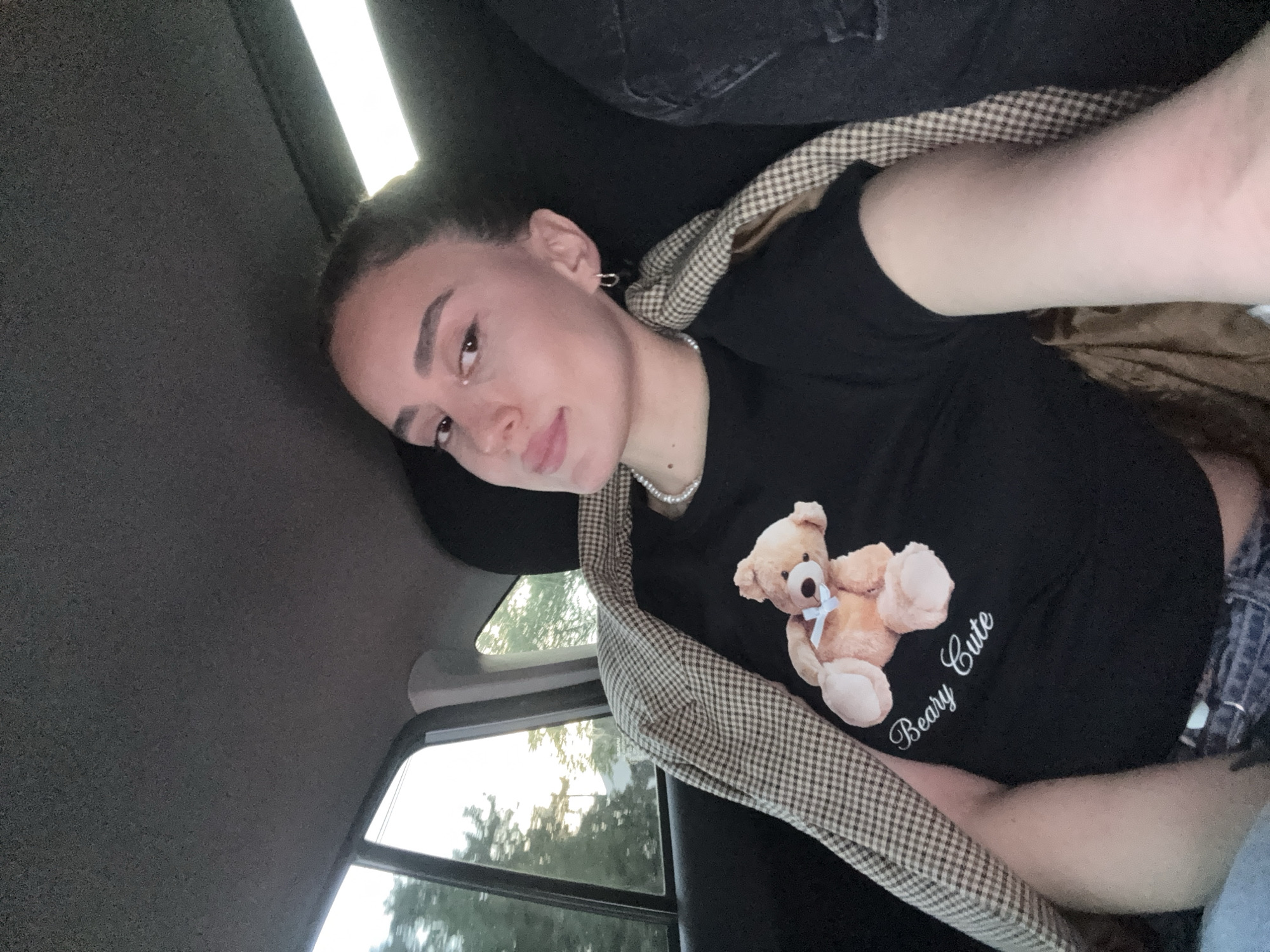 A young woman is sitting in a car, possibly in the city of Tbilisi, Georgia. The woman is facing the camera with her head resting against the headrest. She has light skin, dark hair pulled back, and is wearing a black shirt with a teddy bear graphic that reads "Beary Cute". A checkered jacket or blanket is draped around her shoulders. A car window is visible, showing trees and a hint of the outside environment. The car's interior is mostly dark, with a grey headliner and black seating.