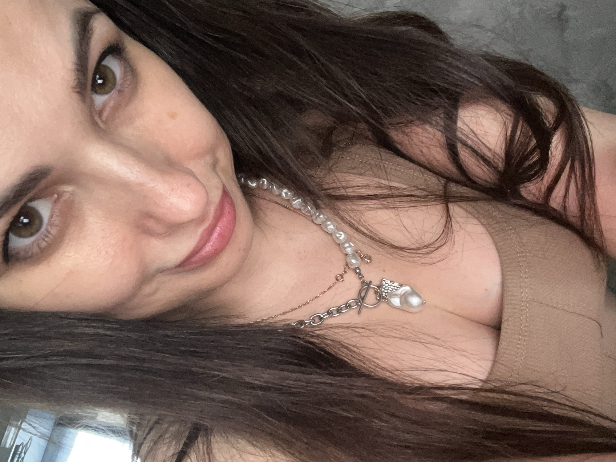 A close-up, tilted portrait features a woman with long, dark brown hair and warm brown/hazel eyes, looking directly at the viewer. Her head is angled to the right, and she has a gentle, inviting smile on her pinkish-mauve lips. Eyeliner is visible on her upper eyelids.
She is wearing a necklace that combines pearls with a silver chain, featuring a toggle clasp and a distinctive baroque pearl pendant. The pearls appear slightly irregular in shape. Her clothing consists of a light brown or beige ribbed top, possibly a tank top or camisole, visible around her upper chest.
Her dark hair frames her face and cascades around her shoulders and chest. The background appears to be an indistinct, light grey surface, suggesting an indoor setting. The lighting is soft and even, highlighting her facial features. No specific time of day or location cues are apparent. There is no visible text in the image.
