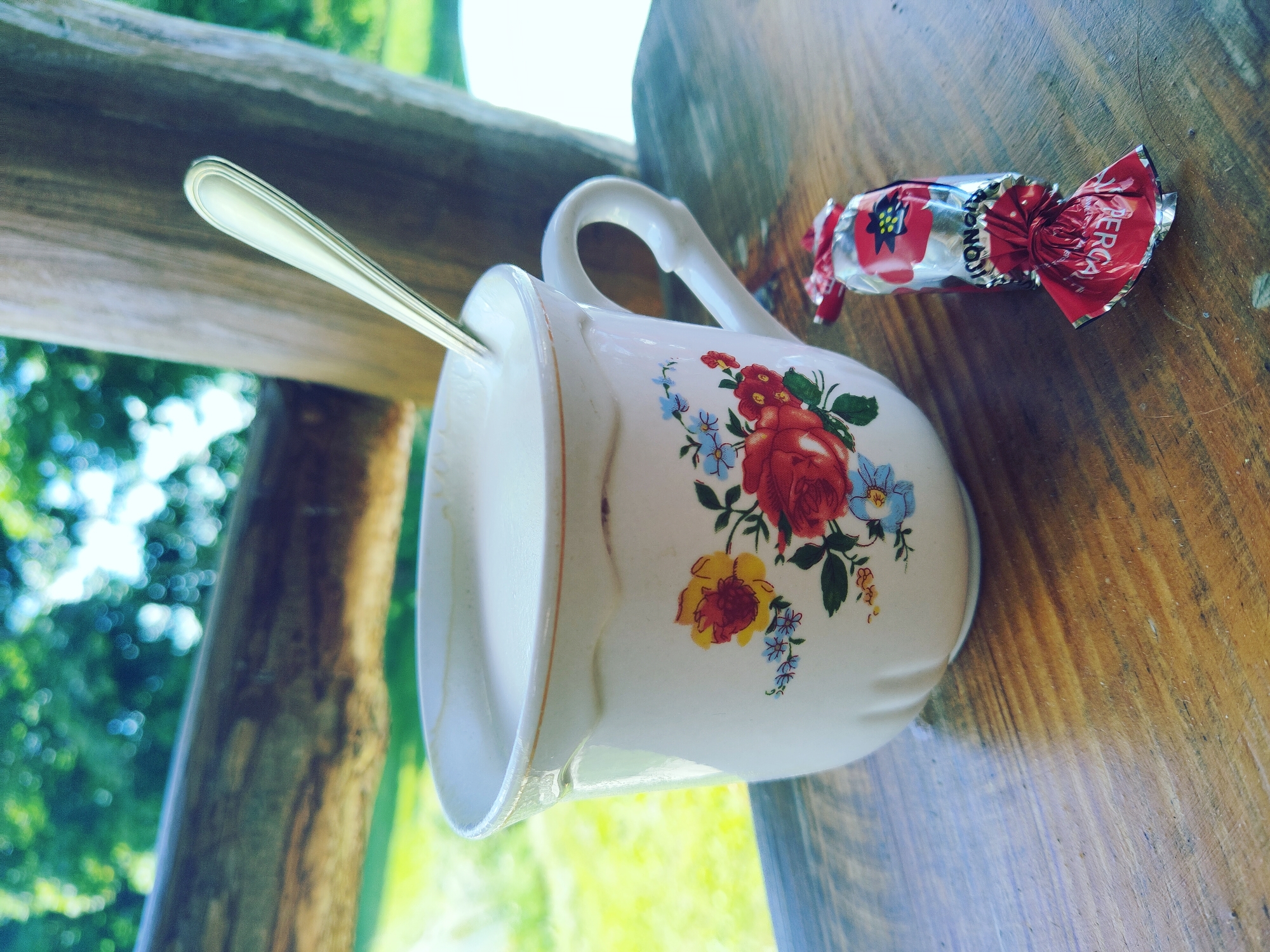 A close-up shot features a white ceramic mug with a floral design, filled with a light-colored liquid, likely coffee or milk. A silver spoon rests in the mug. Next to the mug, on a wooden table, lies a red and silver candy wrapper. The background is blurred, showing greenery and what appears to be a wooden structure, suggesting an outdoor setting. The scene is likely located in Norviliškės, Lithuania.