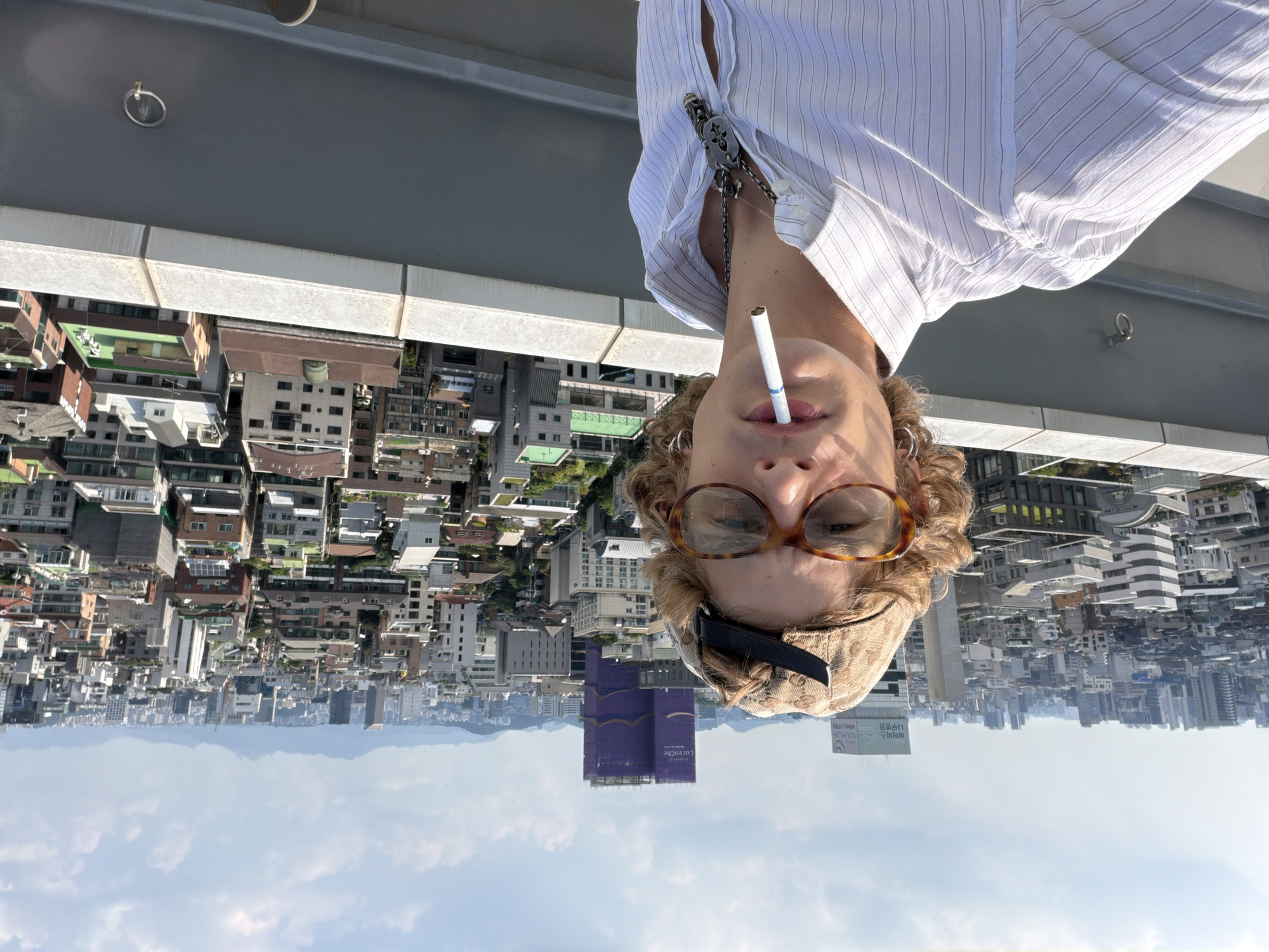 A young man with curly brown hair, wearing a baseball cap, glasses with tortoiseshell frames, and a pinstriped collared shirt, is taking a selfie on a rooftop. He has a cigarette in his mouth and is wearing a necklace. The background shows a sprawling cityscape of Seoul, South Korea, with numerous buildings of varying heights and architectural styles under a cloudy sky. In the distance, mountains are visible. The overall impression is a casual portrait taken in an urban environment.