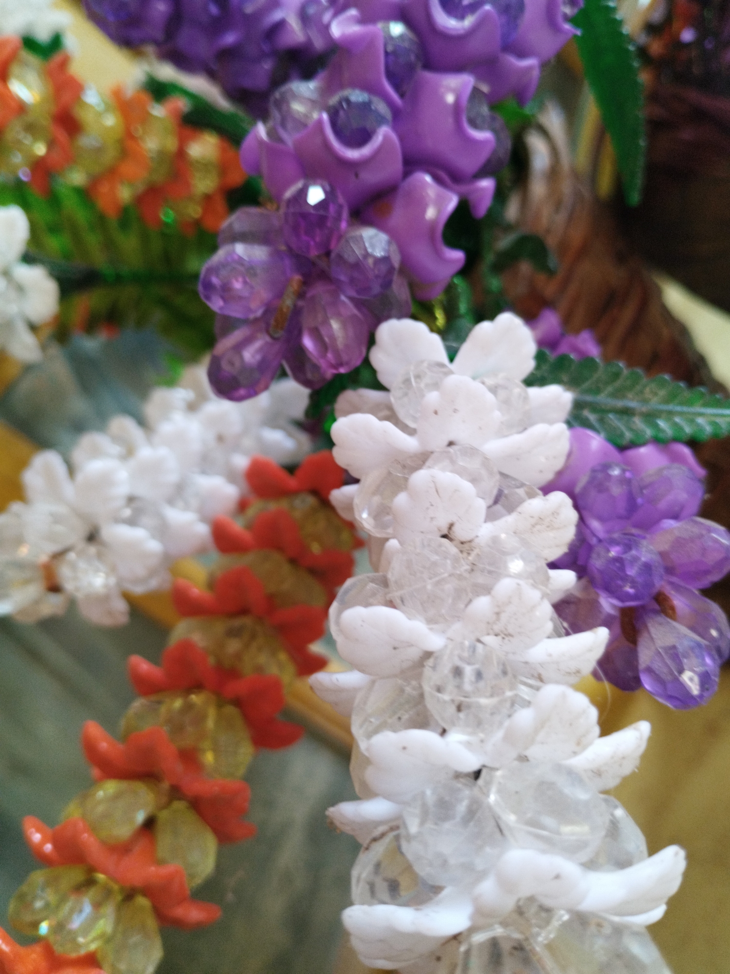 The image shows artificial flowers of different colors, including purple, white, and orange, with a possible location in Lhokseumawe, Indonesia. The flowers are made of beads and arranged in a cluster.