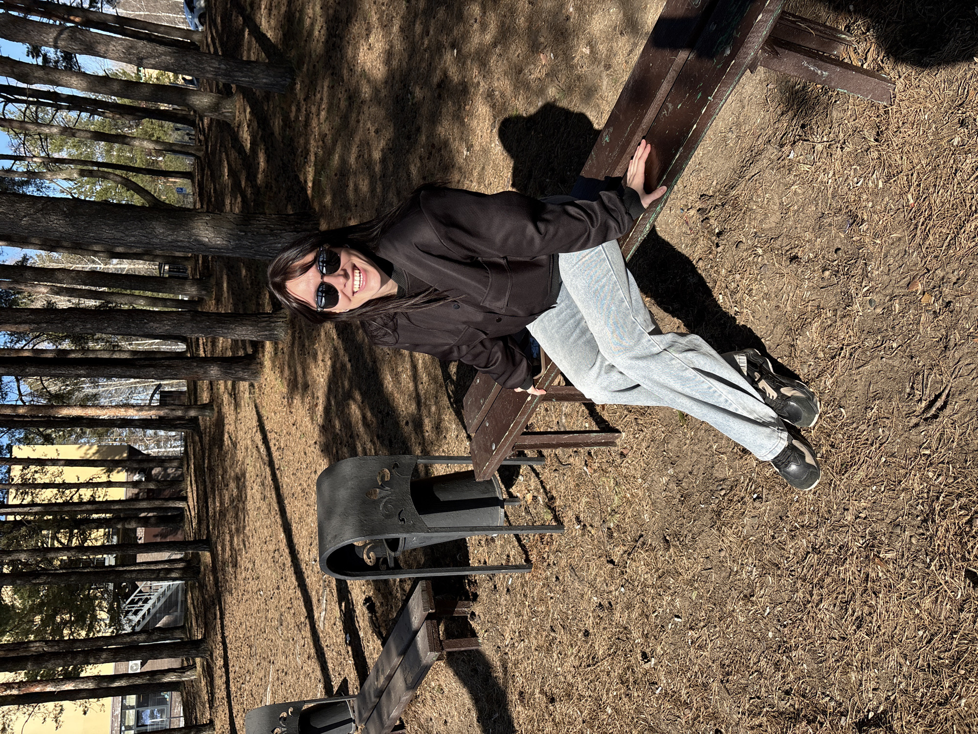 A young woman is captured in a sunny outdoor setting, appearing to sit on a wooden bench in what seems to be a park or wooded area, possibly in Chaikovsky, Russia. She is smiling broadly and wearing dark sunglasses, a dark long-sleeved jacket, light blue jeans, and dark sneakers. Her left hand rests on the back of the bench, suggesting a relaxed pose for the camera.
The scene is dominated by numerous tall, slender pine trees, casting long shadows across the dry, brown ground, which is covered with pine needles and sparse dry grass. The bright sunlight indicates a clear day, likely late morning or early afternoon. In the background, beyond the trees, glimpses of buildings are visible, including a yellow-colored structure and another building with an external metal staircase, suggesting an urban or suburban edge to this natural space. To the left of the woman, a decorative black metal bench or chair with ornate details is also visible. There is no visible text in the image.