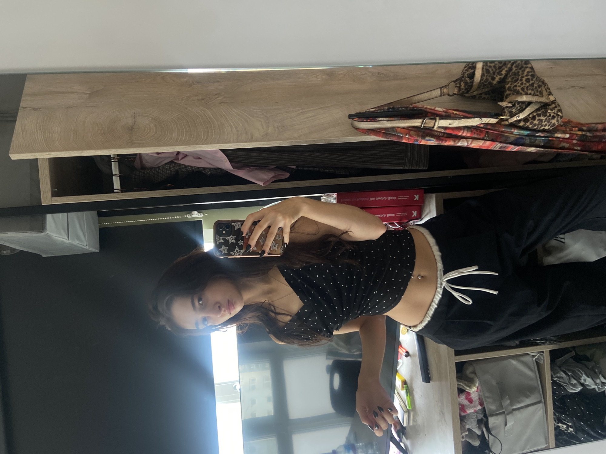 A young female, appearing to be in her late teens or early twenties, stands reflected in a full-length mirror, holding a smartphone in her right hand to take a mirror selfie. She has long, dark hair and wears a black polka-dot spaghetti-strap crop top, black wide-leg trousers with a white drawstring waistband, and a silver belly button piercing. Her fingernails are painted dark. Her left hand is positioned near a desk, holding a thin, dark object.
The background, visible in the reflection, depicts an indoor setting within the city of London, United Kingdom. To the left of the subject, a desk or table holds various items including a blue plastic bottle, a Marlboro Gold cigarette pack, a lighter, multiple pens and markers, and a book spine partially visible, reflecting "London Review of Books." Behind the desk is a black office chair and a window with a lowered black roller blind, revealing urban buildings outside. To the right of the subject, an open wooden cabinet with a light wood grain finish displays clothing on hangers, including a pink shirt and a striped garment. A multi-colored patterned scarf and a leopard-print shoulder bag with light straps hang near or on the cabinet door. The wall to the far right is white.