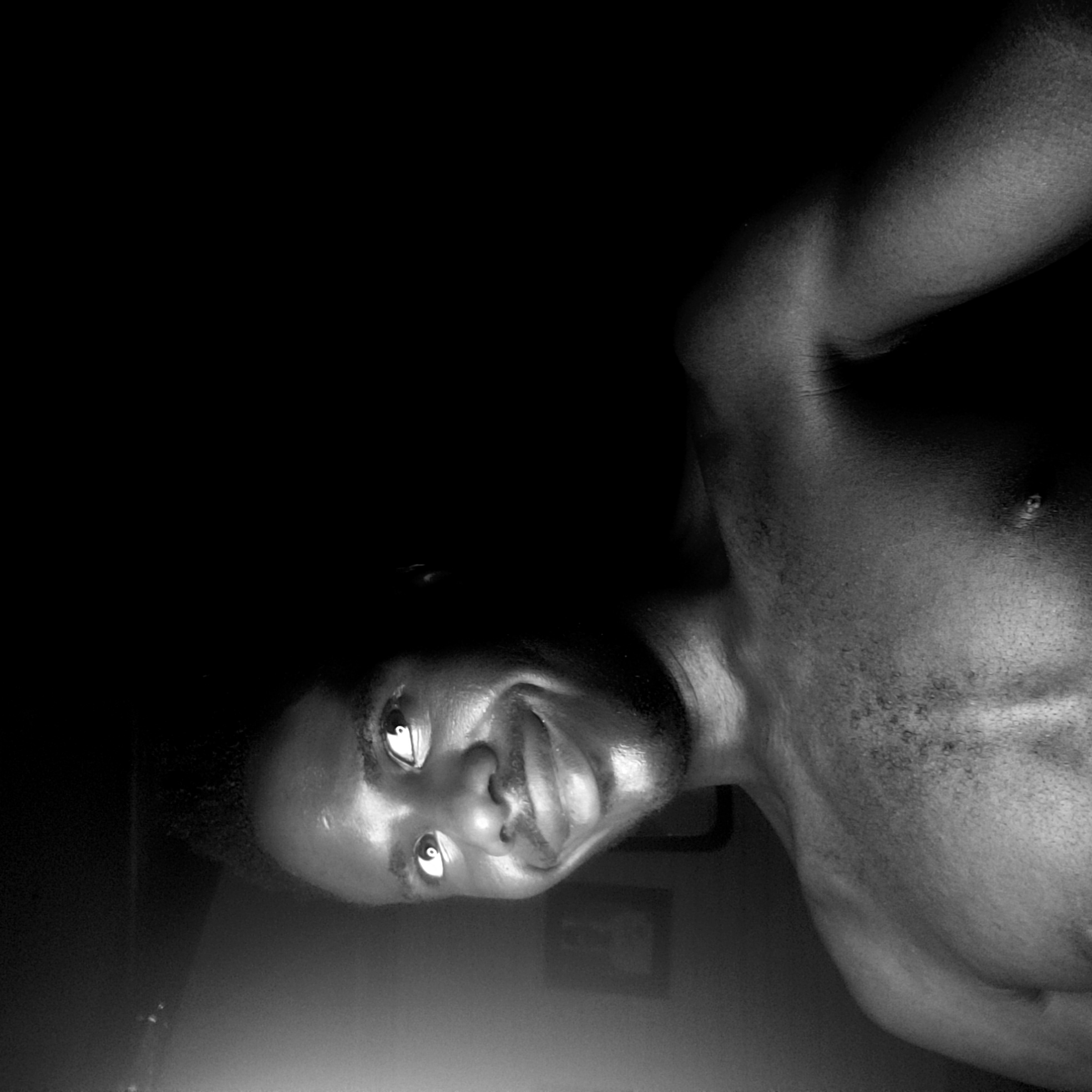 A black and white low-angle photograph shows a shirtless man with dark skin. He has short, dark hair and a slight smile. His eyes are wide open and reflecting light. The background is mostly dark. A partially visible picture frame is on the wall behind him. The location is Narayi, Nigeria.