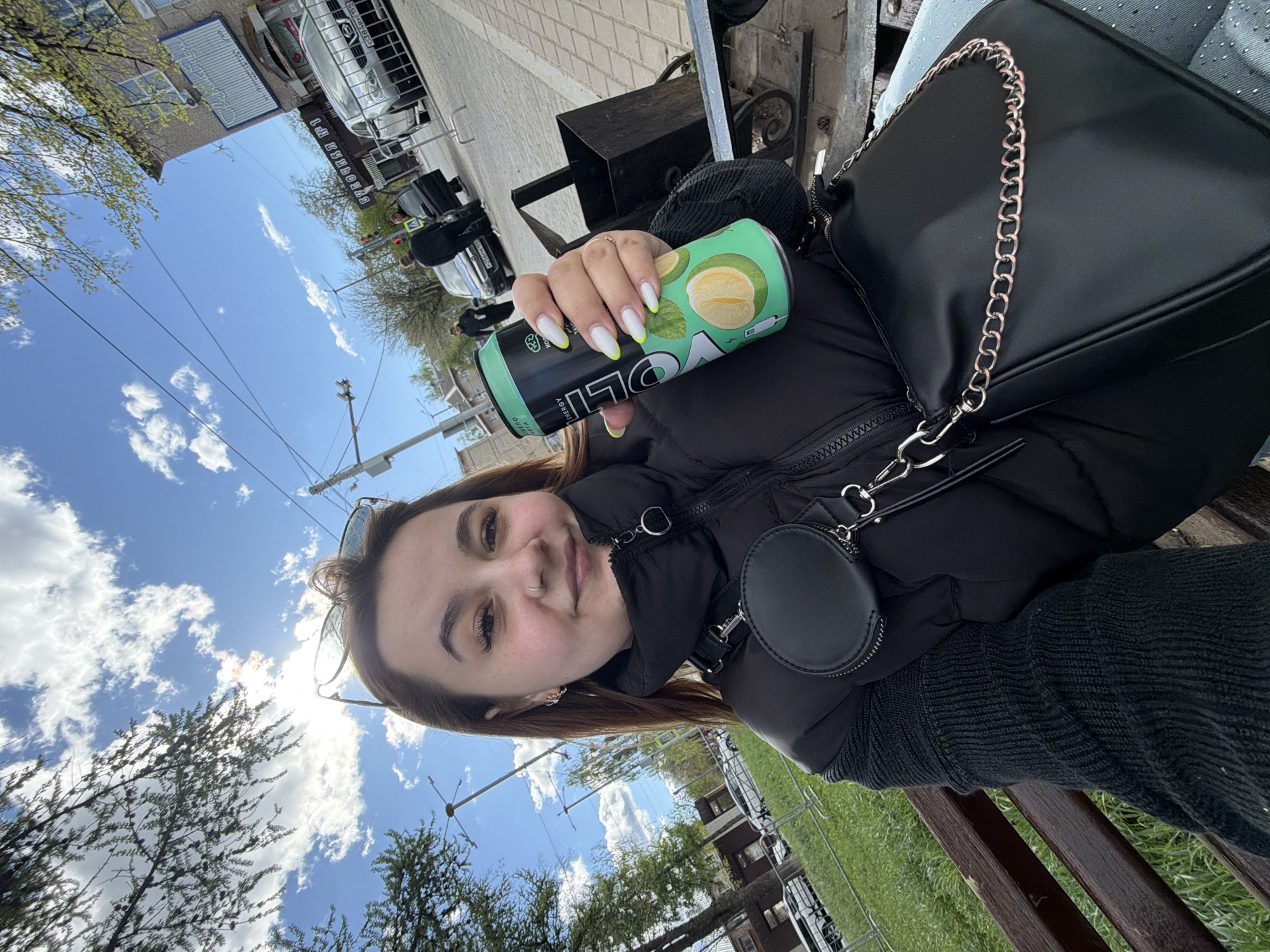 A young woman is seated outside, possibly on a bench, and is holding a can of "VOLT Energy" drink. She's wearing a black puffer jacket, a black sweater, and a black purse with a chain strap. The woman has long nails that are painted white with a neon green accent at the tips.
The background shows a street scene in Chaikovsky, Russia. There are buildings, trees, parked cars, and a blue sky with clouds.