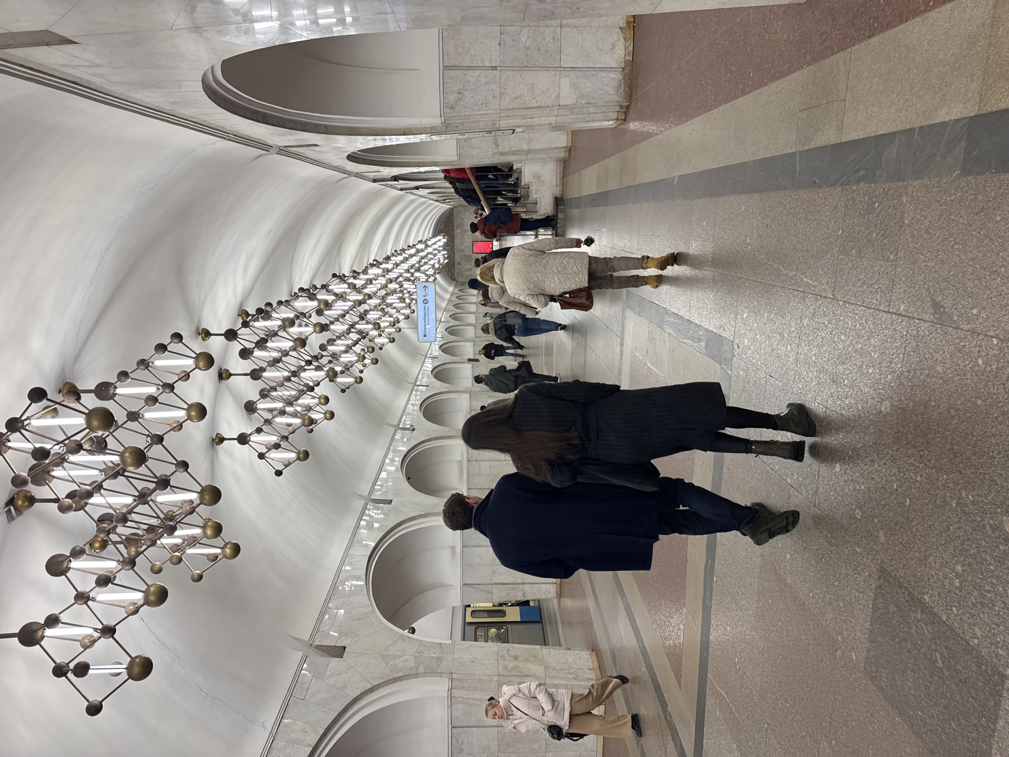 The image depicts a subway station in Khimki, Russia. The architecture includes a curved, white ceiling with arched supports along the walls. There are modern chandeliers hanging from the ceiling. People are walking along the platform. A couple is walking in the foreground. A subway train can be seen through an opening in the wall. The floor is made of granite tiles in different colors and patterns.
