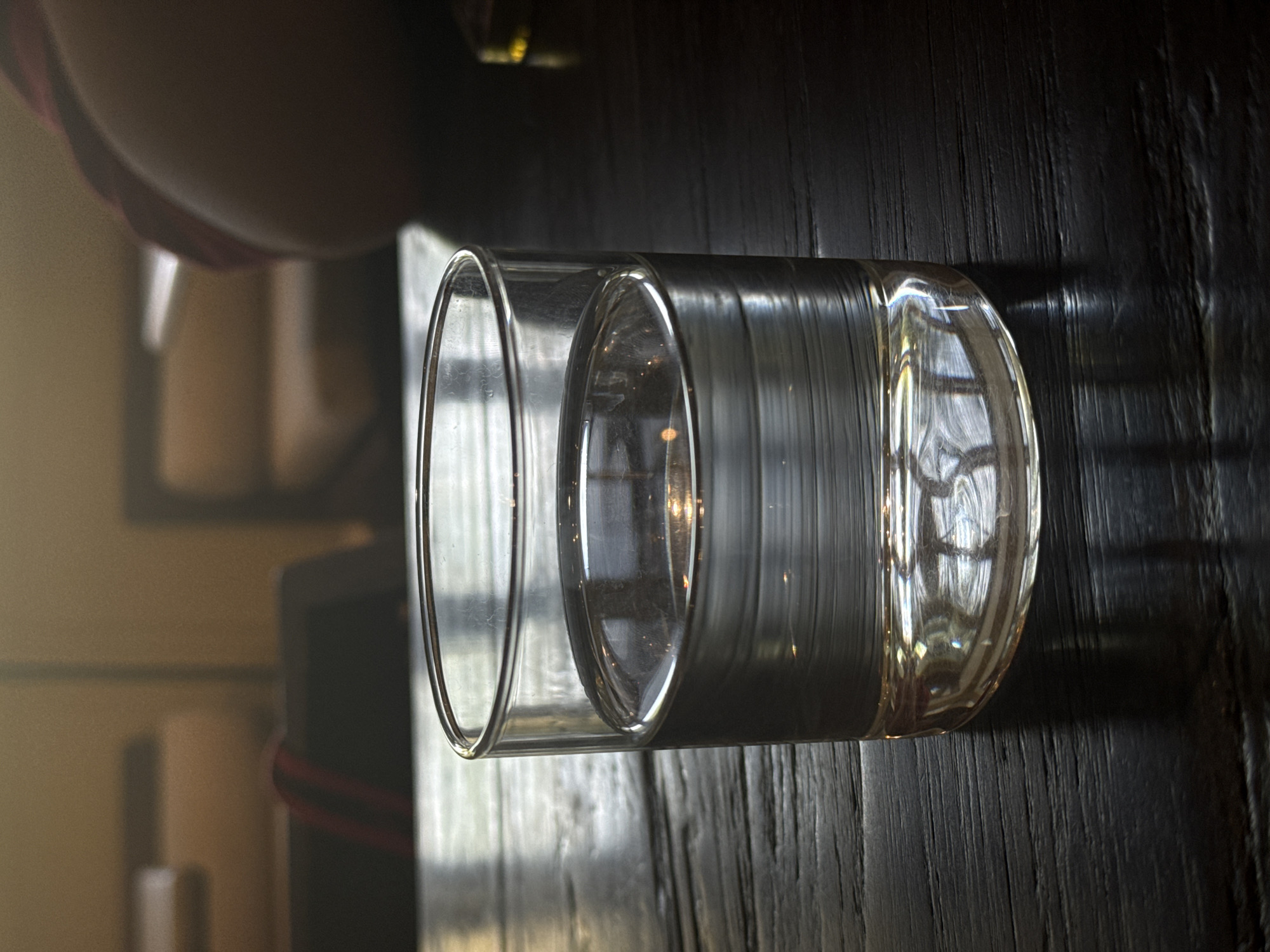 A clear, empty, double-walled glass tumbler rests on a dark, textured wooden surface in a close-up, still-life composition. The glass is cylindrical, showcasing strong reflections and refractions of light that highlight its smooth, transparent material and subtly ribbed appearance. The dark wooden surface, possibly a tabletop, displays distinct vertical grain patterns and catches some ambient light.
The background is softly blurred, creating a bokeh effect. It features indistinct shapes and varying shades of light, with a prominent reddish element visible to the left, which appears to be part of a strap or fabric, possibly belonging to a bag or piece of furniture. The lighting suggests natural light sources, creating bright highlights on the glass and casting soft shadows. There are no people visible, and no specific activity is taking place. The overall mood is calm and contemplative. No text is visible in the image. While the location is North Kuta, Indonesia, no specific geographical cues are present within the frame.