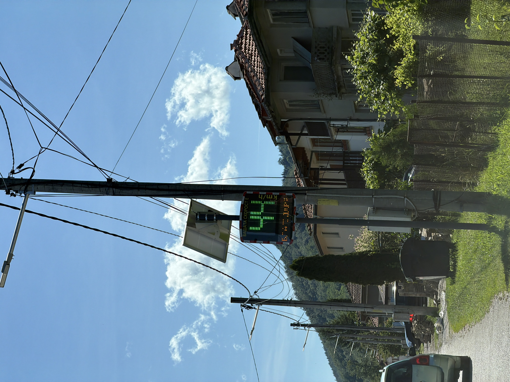The image depicts a street scene in Beli Osam, Bulgaria, under a bright blue sky with scattered white clouds. A speed indicator mounted on a utility pole displays "44 km/h" in green LED digits, preceded by text that appears to read "ВАШАТА СКОРОСТ Е" (Your speed is). Below the speed, there is further text in Cyrillic characters, likely indicating a speed limit or a message. A solar panel is attached to the top of the pole, powering the device.
To the right of the speed indicator, a section of a building with a red tiled roof and weathered, light-colored walls is visible. The building features multiple windows and balconies, one with an ornate railing. A rusty metal fence runs along the property line, separating it from the street.
On the left side of the image, a light-colored car is partially visible, driving away from the viewer on an asphalt road. The roadside is lined with green grass and more utility poles with overhead wires. In the background, a dense green forest covers the rolling hills. A tall, dark green conifer tree stands near the speed indicator, and a large dark trash receptacle is located on the grassy verge. The overall environment suggests a rural or semi-rural setting.