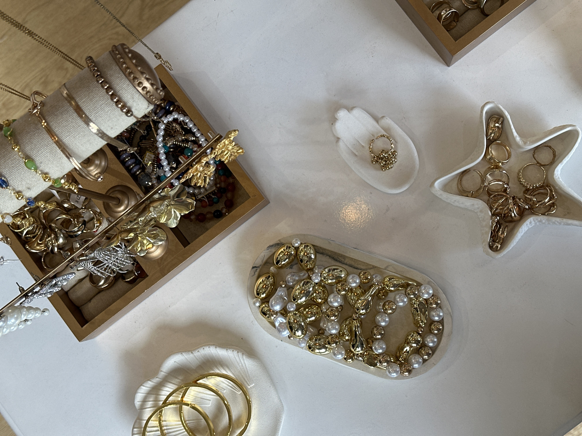 The image shows a tabletop display of jewelry, likely in a shop or market setting. The location could be North Kuta, Indonesia, but this cannot be confirmed from the image alone.
**Objects:**
* Wooden jewelry display stand with various bracelets, necklaces, and earrings.
* Small white ceramic hand-shaped dish holding a gold ring.
* White ceramic starfish-shaped dish filled with gold rings.
* Oval-shaped dish with gold and pearl jewelry.
* White seashell-shaped dish containing gold hoop earrings.
**Environment:**
* The setting is indoors, with a light-colored surface (possibly a table or counter).
* The lighting appears to be bright and natural.
**Subjects:**
* The primary subject is jewelry, including rings, bracelets, necklaces, and earrings.