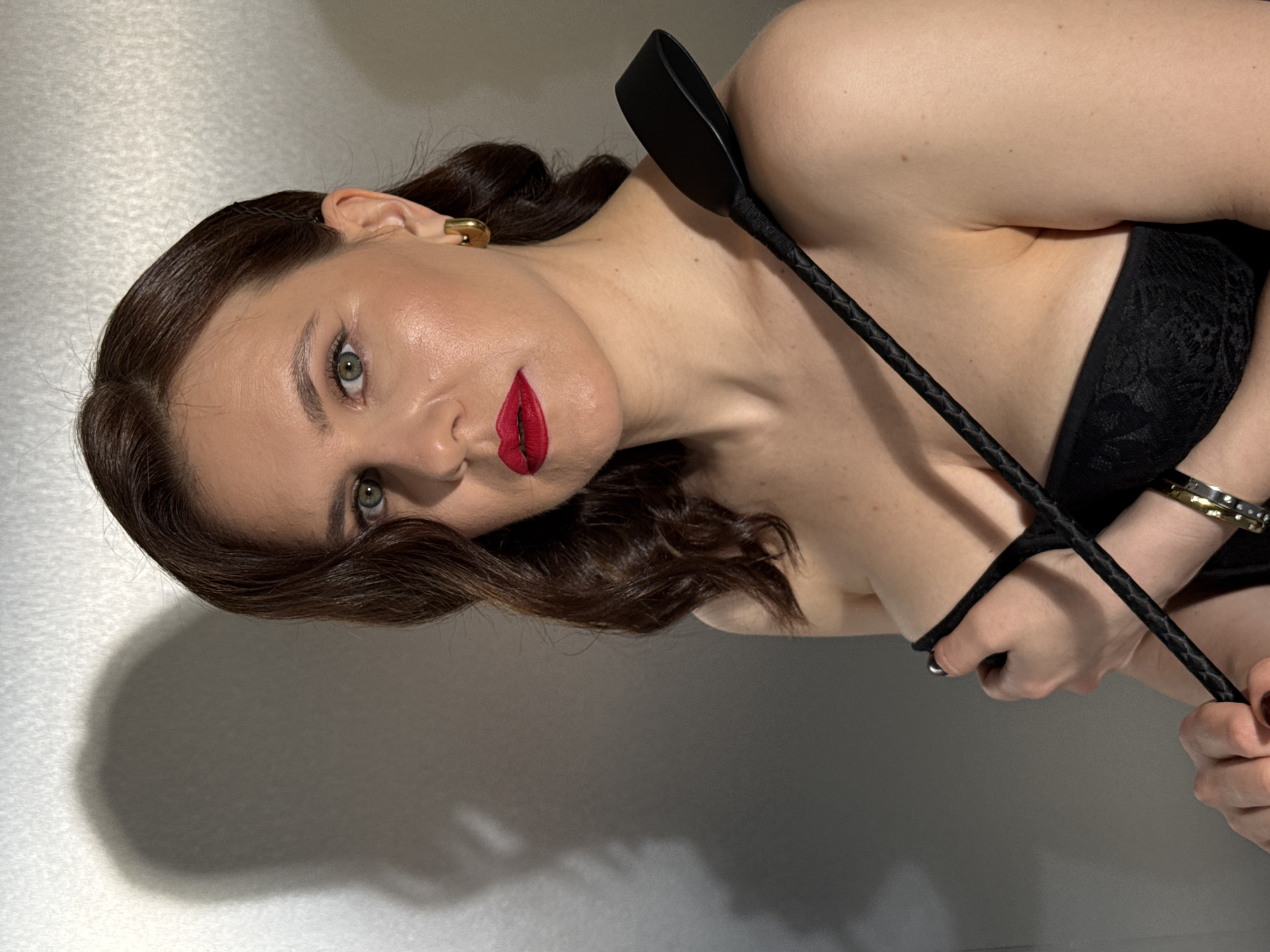 A woman with brown hair and red lipstick is lying down. She is wearing a black bra and holding a black riding crop. She has a gold earring in her left ear and two bracelets on her left wrist. The background is a plain grey wall. There is no indication of the location being Izhevsk, Russia.