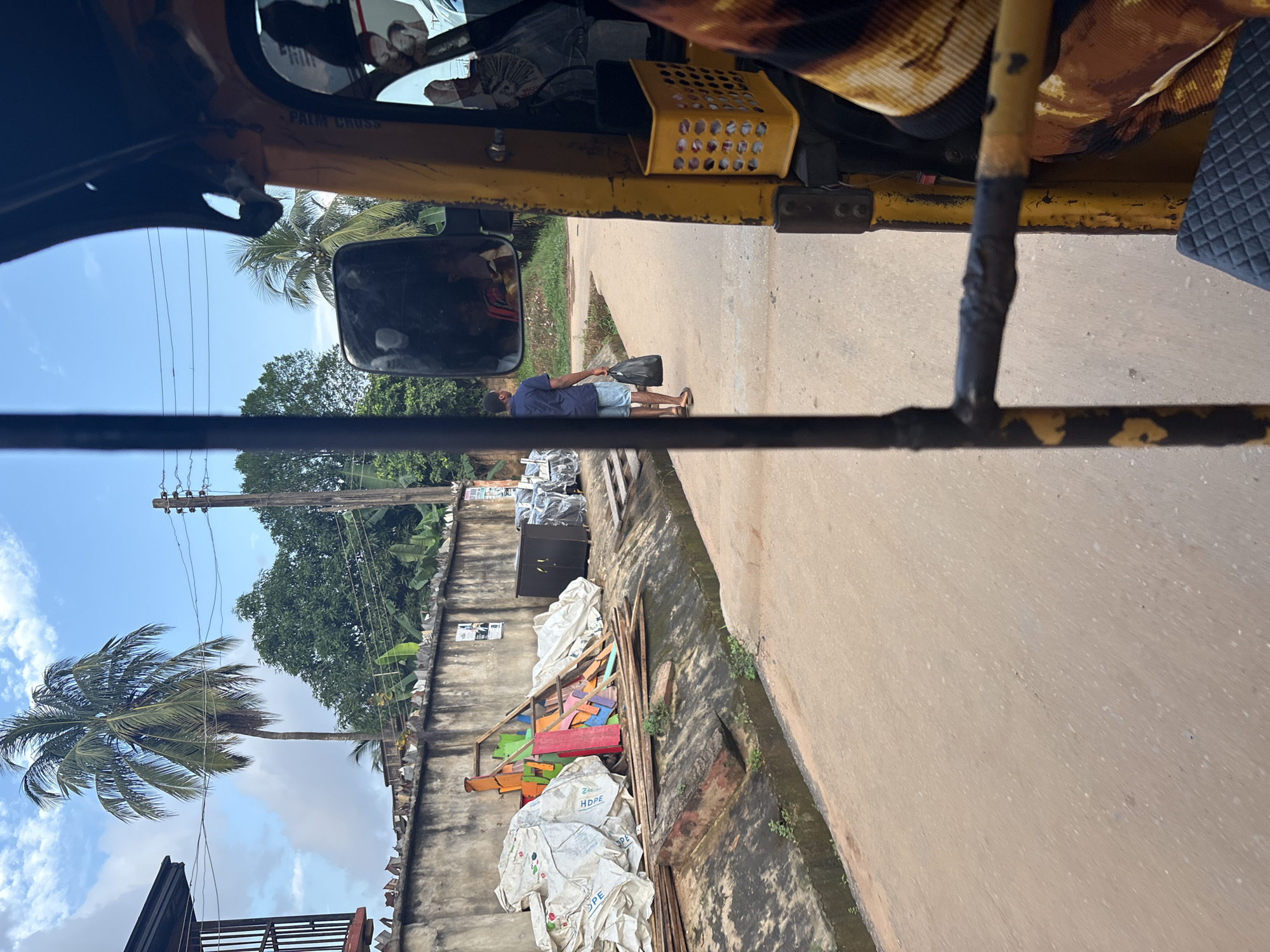 A man in a blue short-sleeved shirt and blue shorts walks away from the camera down a paved street in Onitsha, Nigeria. He carries a black plastic bag in his right hand. He is walking on the right side of the street, heading away from a concrete wall on his left. The wall is topped with pointed debris and has posters affixed to it. To the left of the wall are large white sacks, some colorful wooden planks, and a dark cabinet. A narrow, overgrown drainage ditch runs alongside the wall. In the foreground, the yellow metal frame of a tuk-tuk is visible, obscuring the right side of the frame. A yellow basket is attached to the tuk-tuk. The sky is blue with scattered clouds, and palm trees and other vegetation are visible in the background.