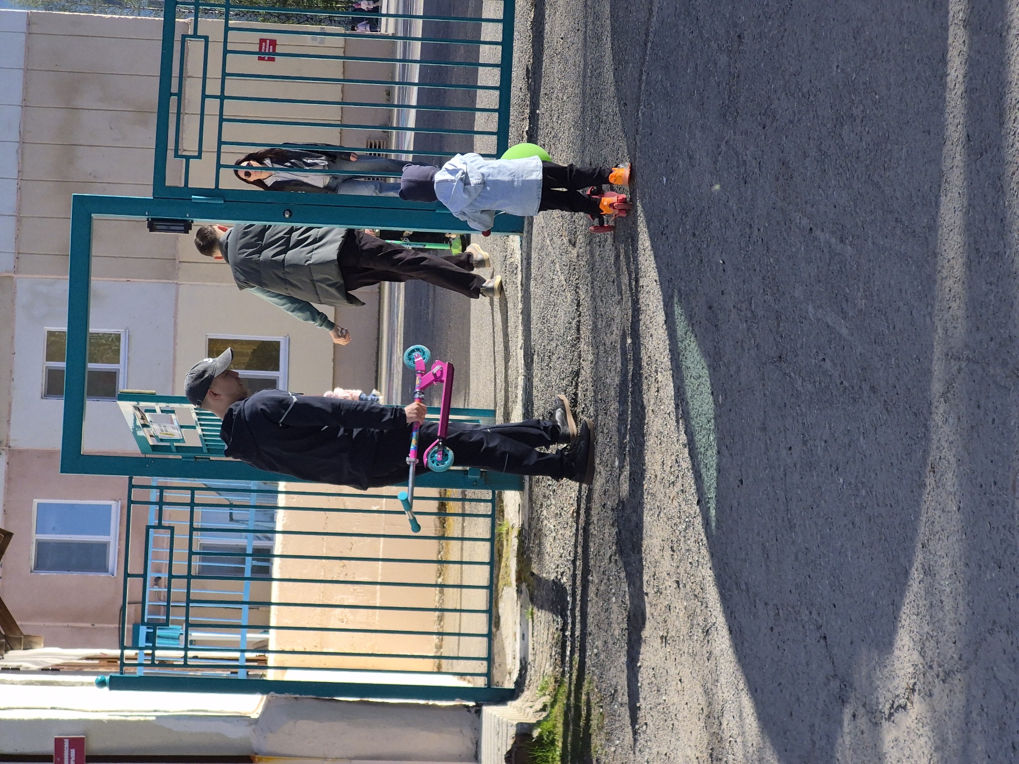 A man in dark clothing and a baseball cap holds a pink and blue scooter near a teal metal gate, adjacent to a beige building with blue-framed windows. Beyond the gate, another man in a green jacket stands next to a child wearing a light blue jacket and roller skates, who holds a green balloon. The scene is on a paved surface, possibly a sidewalk, and is illuminated by sunlight casting shadows. The location is possibly in Tobolsk, Russia, suggested by the architectural style of the buildings.