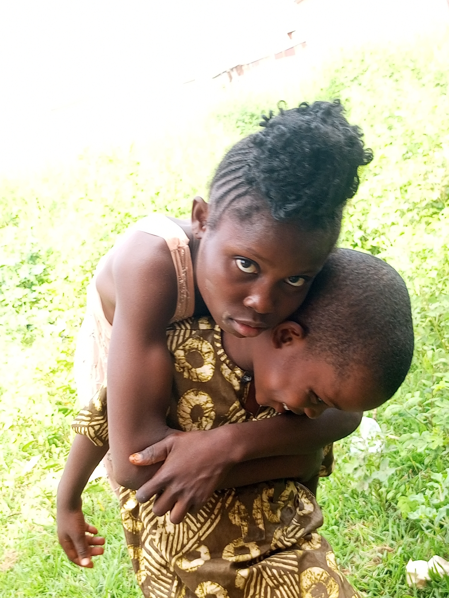 Two young children of African descent are depicted from a high angle, outdoors, with lush green grass and foliage in the background. The child in the foreground, positioned on the viewer's left, is being carried piggyback by the other child. The carrying child, facing away from the viewer, has short, dark hair and is wearing a brown and tan patterned garment with circular designs. The child being carried, facing the viewer, has dark skin and intricate braided hair styled upwards. This child is wearing a light-colored sleeveless top. The interaction between the children suggests a moment of comfort or play, with the carrying child's arms wrapped around the other's torso, and the carried child resting their head on the carrier's shoulder. The lighting indicates it is daytime. The scene is set in a natural outdoor environment, consistent with a rural or semi-rural location, possibly in Tempure, Nigeria.