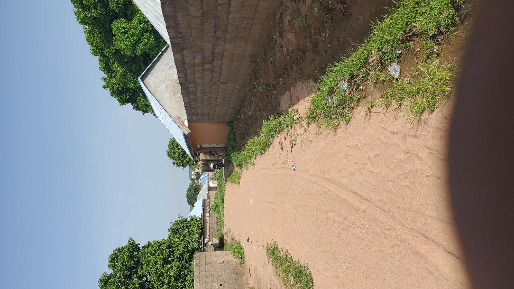 The image depicts a dusty, unpaved street in Jalingo, Nigeria. The street is bordered by a low, grey concrete block wall on the right, behind which is a building with an orange facade and a light blue metal roof. Further down the street on the right, another building with a grey corrugated metal roof is visible. On the left side of the street, there is a concrete block wall, and beyond that, trees with dense green foliage. Several smaller buildings with light blue metal roofs are visible in the distance on the left. A person is riding a motorcycle down the street in the mid-distance, heading away from the camera. There is some scattered litter on the side of the road and on the street itself. The sky is clear and pale blue. The foreground shows a shadow of a person's hand, indicating the photographer's presence. The overall impression is of a semi-urban or rural setting in a warm climate.