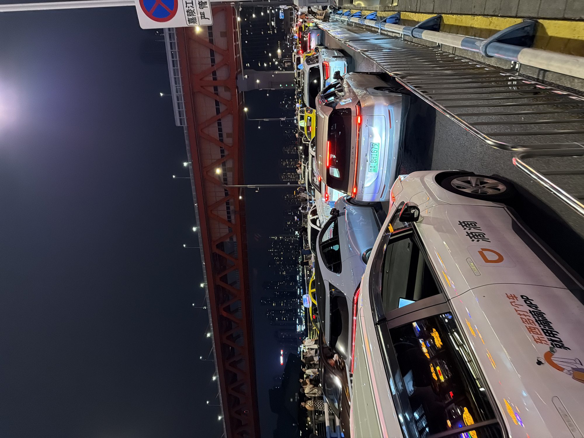 The image captures a traffic jam on a multi-lane road at night, likely on a bridge or elevated roadway in the Yuzhong District of Chongqing, China. A prominent orange bridge structure spans across the upper portion of the frame, with its supporting trusses clearly visible. The sky is dark, with some lights from distant buildings forming a cityscape silhouette in the background.
On the road, numerous vehicles are stalled, indicating heavy congestion. Several cars are white, with some bearing the distinctive orange logo and Chinese characters of the ride-hailing service "Didi." One white Didi car in the foreground displays a license plate with characters "A BH1679." Other vehicles in the traffic include silver SUVs, a yellow taxi, and darker colored cars. Headlights and taillights are illuminated, creating streaks of light in the darkness.
Along the right side of the road, a metal barrier separates the traffic lanes from a sidewalk or walkway. A traffic sign with a red circle and a diagonal blue bar is visible on a pole next to the barrier, indicating a "no stopping" or "no parking" restriction. The road surface appears wet, reflecting the vehicle lights.
On the left side of the frame, a group of people are standing near the edge of the elevated road, looking out at the traffic or the city. They are dressed in casual evening wear.
The overall impression is one of urban nighttime activity, characterized by heavy traffic and infrastructure characteristic of a major Chinese city.