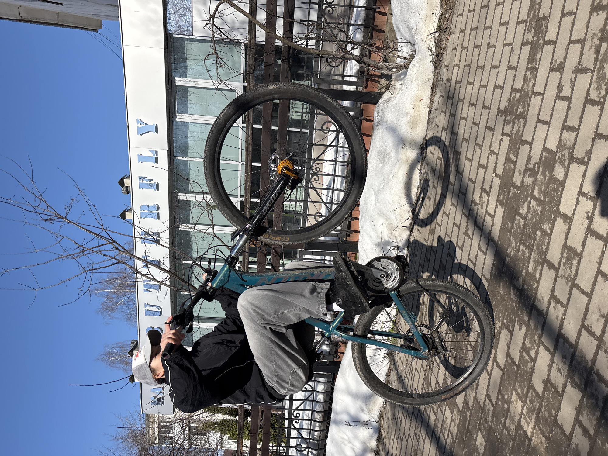 A young man is performing a wheelie on a mountain bike on a brick sidewalk in Chaikovsky, Russia. He is wearing a black jacket, gray pants, and a gray and white baseball cap. The front wheel of the bike is raised in the air, and the rear wheel is on the ground. The background includes a building with a sign that reads "ТЕАТР". There is snow on the ground near a metal fence. The sky is blue and sunny, and the shadow of the bicycle is visible on the brick sidewalk.