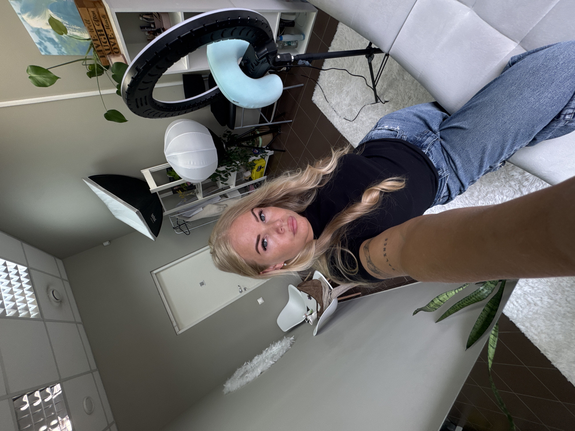 A woman with blonde hair is lying on a white sofa, wearing a black shirt and blue jeans. A ring light and a softbox light fixture are positioned nearby. A plant and a painting are placed on shelves in the background. The location appears to be indoors, with a white ceiling and a neutral-colored wall.