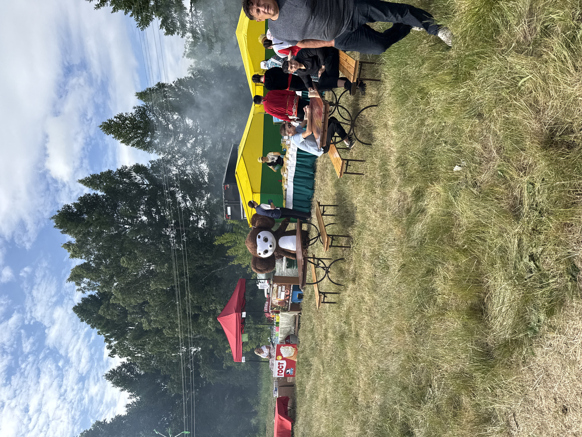This image depicts an outdoor fair or festival scene set in a grassy field, possibly in Nurykeevo, Russia, judging by the contextual information. In the foreground, a man in a gray t-shirt and jeans is looking towards the camera. To his left, two men are seated at a wooden picnic table. Behind them, several yellow and green tents are set up, with people milling about and food being served. A large inflatable Cheburashka character stands prominently near the tables. Various other stalls and tables are visible, some covered with red cloths, indicating food vendors or sales booths. The background features a line of tall trees and a partly cloudy sky. Smoke is rising from behind some of the tents, suggesting cooking or a barbecue.