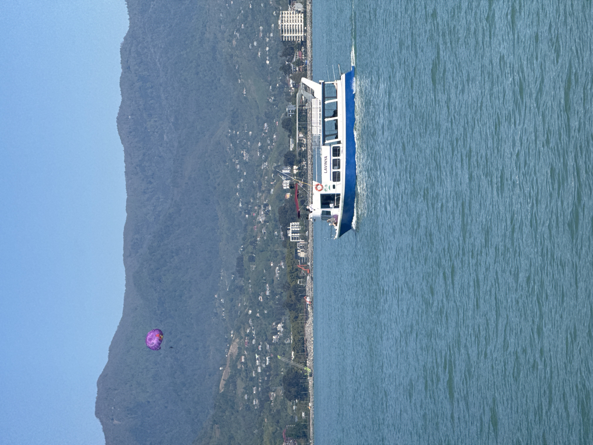 The image depicts a body of water alongside a coastline in Batumi, Georgia. A blue and white vessel, labeled "LAVINYA," is navigating the water. In the background, a mountainous landscape is partially visible. A purple parasail is seen in the sky near the mountainside. The coastline shows infrastructure and buildings extending along the base of the mountain range.