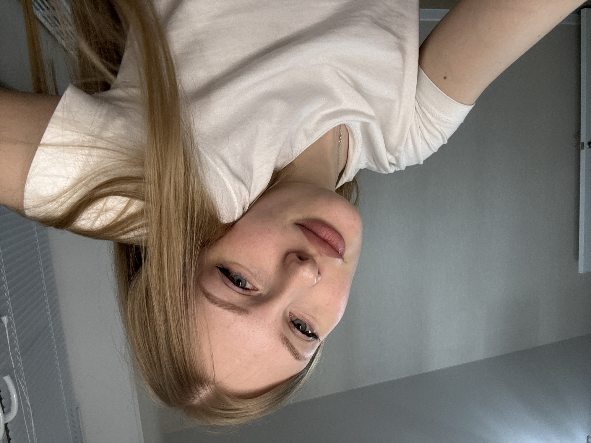 The media file displays a rotated, close-up selfie of a young person, likely female, with fair skin, light brown hair, and green eyes. She is looking directly at the camera with a neutral, slightly casual expression. Her head is tilted, and her mouth is slightly open, revealing a small white mark or substance near her lower lip. She is wearing a plain white t-shirt, and a delicate silver-colored necklace is visible around her neck.
The activity taking place is a self-portrait (selfie). The setting appears to be indoors, against a plain, light-colored wall. On the right side of the frame, a vertical white structural element or door frame is partially visible. To the left, a subtly textured or patterned surface, possibly window blinds or a textured wall, can be discerned. The lighting suggests an indoor, likely daytime, environment. No specific time of day or weather conditions are discernible. There are no visible texts within the image. While the location context is provided as Perm, Russia, there are no visual cues within the image itself to confirm this specific geographical location.