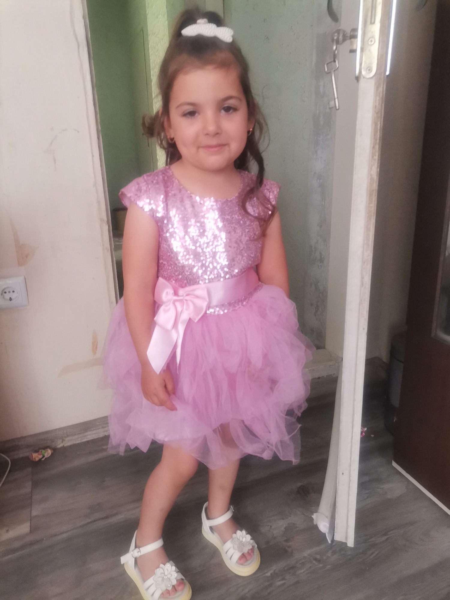 A young girl stands indoors, wearing a pink, sparkly dress with a large pink bow at the waist and white sandals. She has a white hair accessory. The background includes a doorway, an electrical outlet, and a door. The floor is gray. A key is hanging on the door.