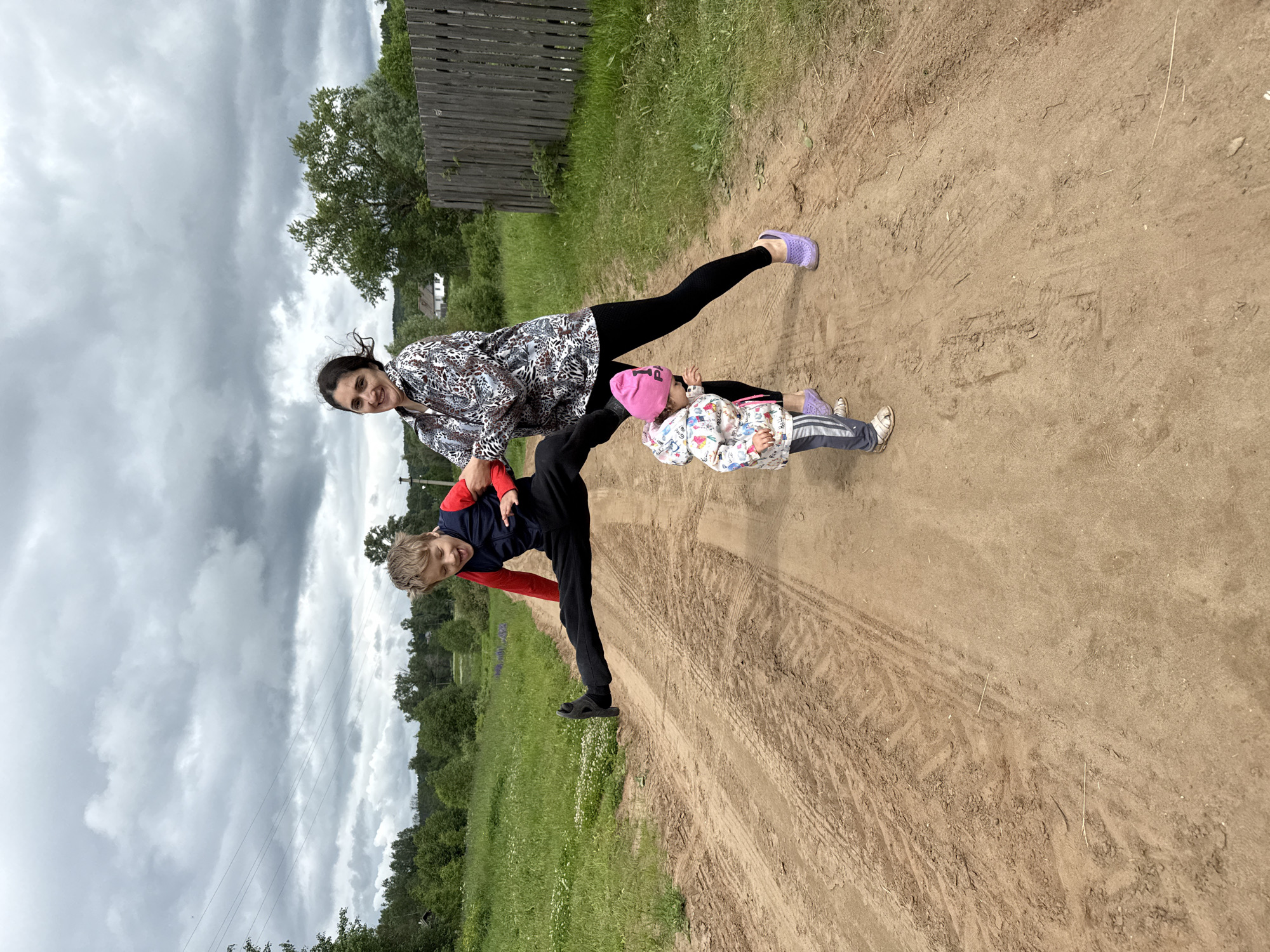 This image depicts three individuals—an adult female, a pre-adolescent boy, and a toddler—engaged in an outdoor activity on a dirt road in a rural setting, identified as Уральское, Russia.
The scene is dominated by an overcast sky with heavy grey cloud cover. In the background, a dense line of green deciduous trees stretches across the horizon, with what appears to be a distant white building and a utility pole visible on the left. A weathered wooden picket fence runs along the right side of the frame, bordering a grassy area.
The foreground is a wide, light brown dirt thoroughfare, deeply rutted with distinct tire treads and scattered footprints. Lush green grass and small white wildflowers line both sides of the road.
The adult female, positioned slightly right of center, wears a long-sleeved, button-up shirt featuring a complex, multi-colored animal print pattern, black leggings, and purple slip-on shoes. She has dark hair, which appears wind-blown, and is smiling directly towards the camera. Her right hand grasps the left arm of the boy. Her stance is active, with knees slightly bent, suggesting she is supporting or assisting the boy.
The boy, in mid-air to the left of the woman, is performing a wide-legged jump, almost a horizontal split, several inches above the dirt road. He wears a long-sleeved red shirt, a dark sleeveless vest, black pants, and dark shoes. His light brown hair is disheveled, and his mouth is open with his tongue slightly extended, indicating an expression of exertion or excitement. He appears to be approximately 8-12 years old.
The toddler stands on the dirt road between the woman's legs and slightly in front of her. The toddler wears a pink knit hat, a multi-colored patterned hooded jacket, grey and white striped pants, and white sandals. The child is looking upwards towards the jumping boy.
The overall interaction suggests a playful, energetic moment, with the adult facilitating the boy's jump while the toddler observes.