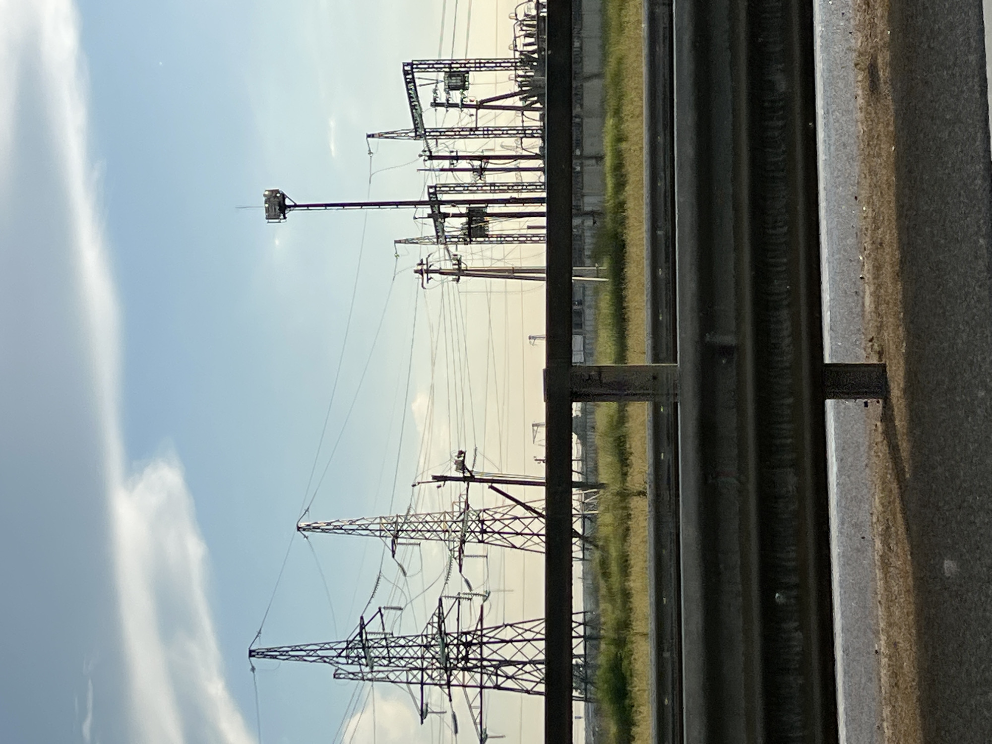 The image depicts an outdoor scene featuring an electrical substation with multiple high-voltage transmission towers and supporting structures. The towers are made of metal lattice and carry numerous electrical wires. In the foreground, there is a guardrail, likely along a road or highway, with a grassy area and a low concrete wall behind it. The sky is partly cloudy with a clear blue expanse. The overall impression is of industrial infrastructure situated in an open area. The image appears to be taken during daylight hours, with the sun casting soft shadows. The location is identified as Psekups, Russia.