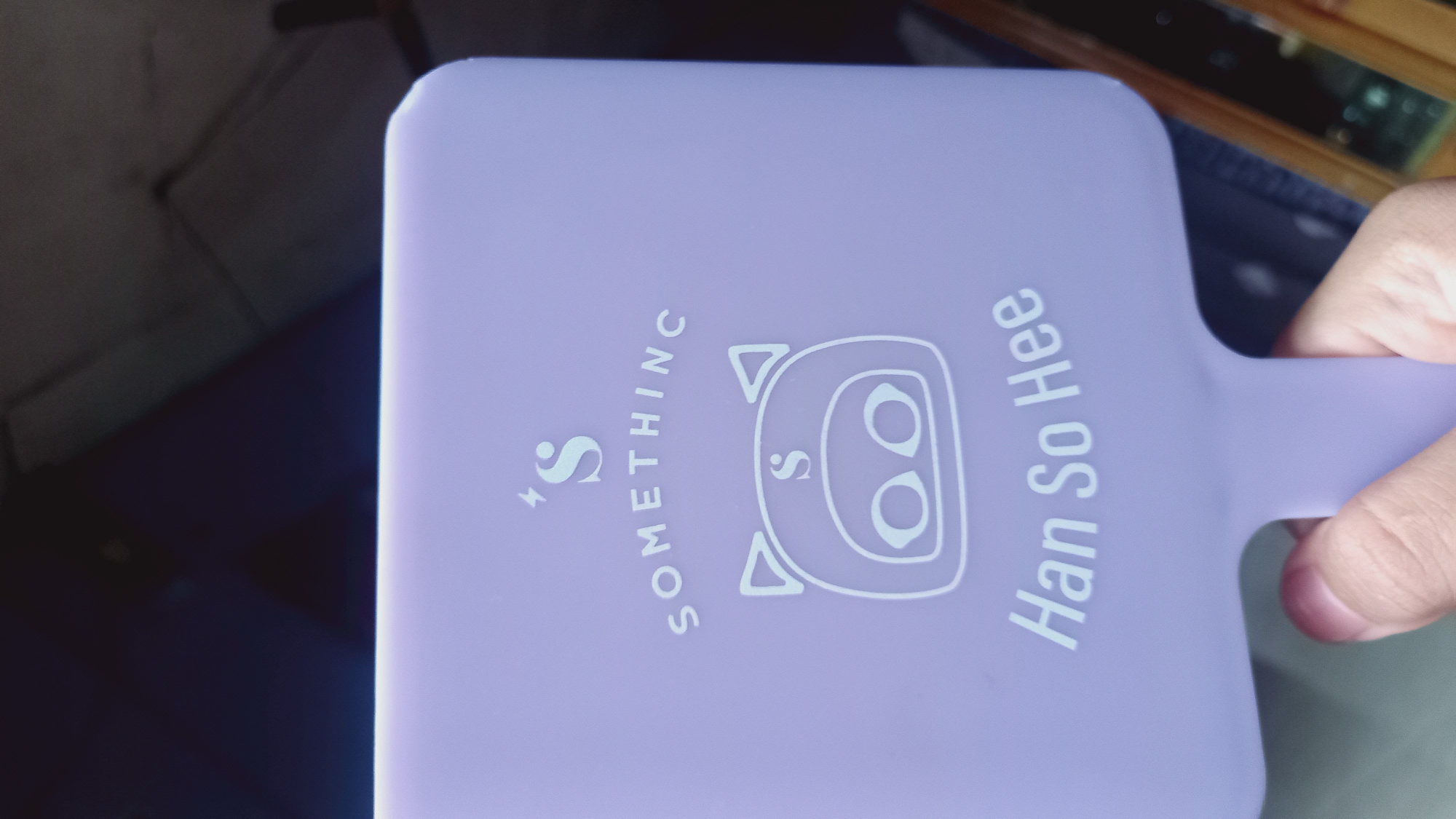 A light purple handheld mirror with a white design is held by a person's fingers. The design features a stylized lightning bolt above the letter 'S', followed by the word "SOMETHING" in an arc. Below this is a simple outline of a pig's face with the letter 'S' inside the pig's forehead and two circles for eyes. At the bottom of the mirror, the words "Han So Hee" are written. The background is dark and out of focus, suggesting an indoor environment. There is no indication of Leuwisadeng, Indonesia in the image.