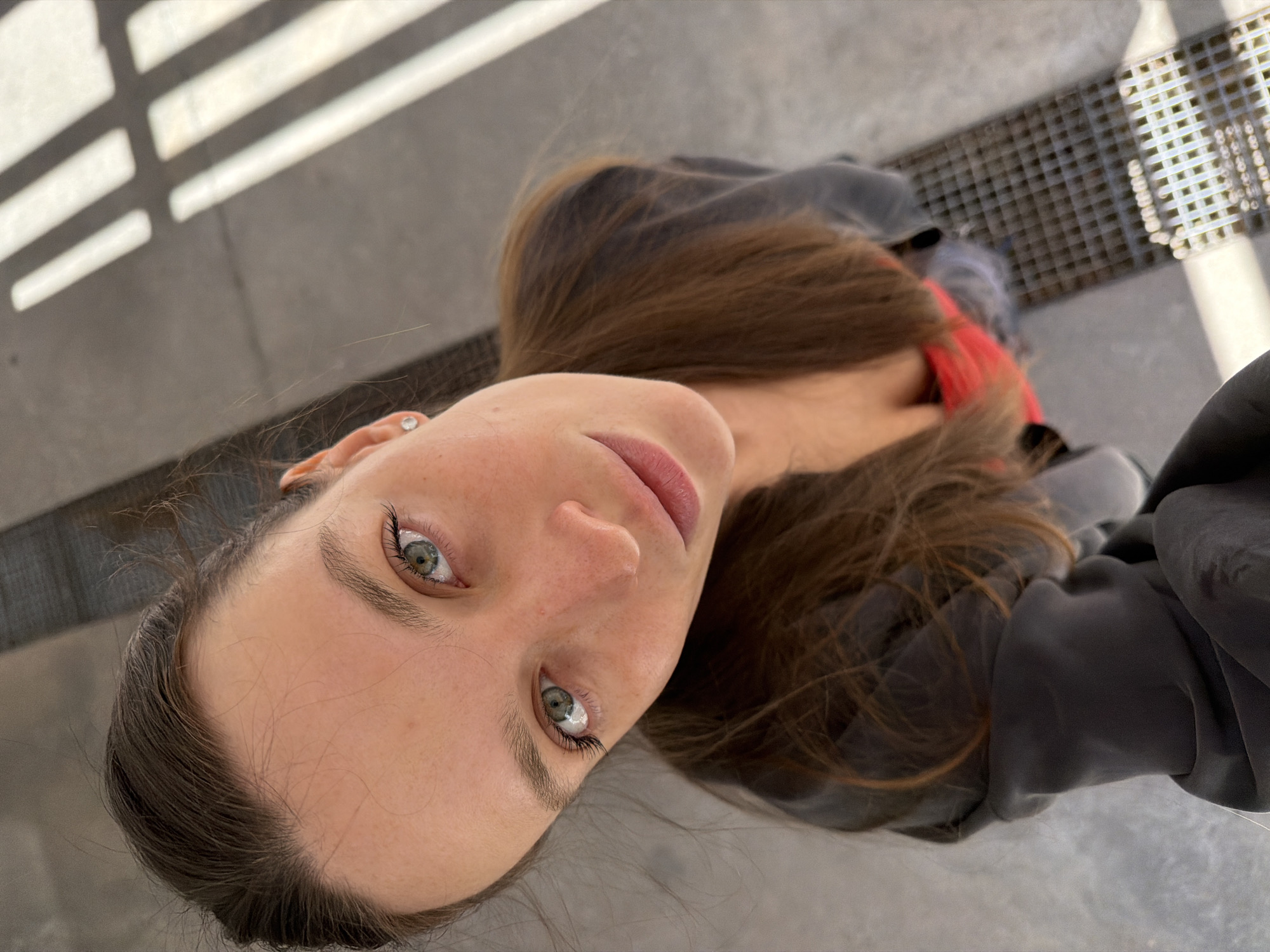 A high-angle close-up portrait features a young woman looking directly upwards at the camera. She has long, straight, reddish-brown hair, and captivating light green or hazel eyes with prominent dark eyelashes, possibly enhanced. Her skin is fair, and her full lips have a subtle pink hue. A small, clear stud earring is visible in her left ear. Her expression is neutral but engaging. She is wearing a dark, possibly grey or black, garment around her shoulders, with a flash of red fabric visible near her neckline, suggesting an inner layer or a detail of her top.
The scene is outdoors on a bright, clear day, likely mid-morning or afternoon, given the sharp, distinct shadows. The background consists of a textured, grey ground surface, possibly concrete or pavement. Strong, parallel shadows, cast by an overhead slatted structure such as a pergola or railing, diagonally stretch across the ground. To the upper right, a metal grate or drain cover is visible on the paved surface. The setting appears to be an urban or architectural outdoor space, consistent with locations found in Budva, Montenegro. No visible text is present in the image.