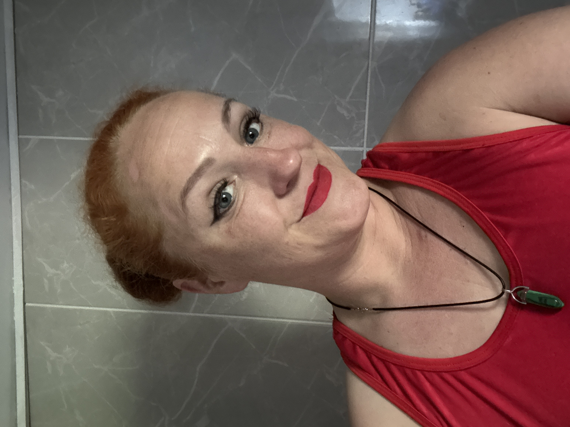 A selfie of a woman in a red tank top, wearing a black necklace with a green stone pendant, positioned in front of a wall with grey tile. The location is not discernible in the image.