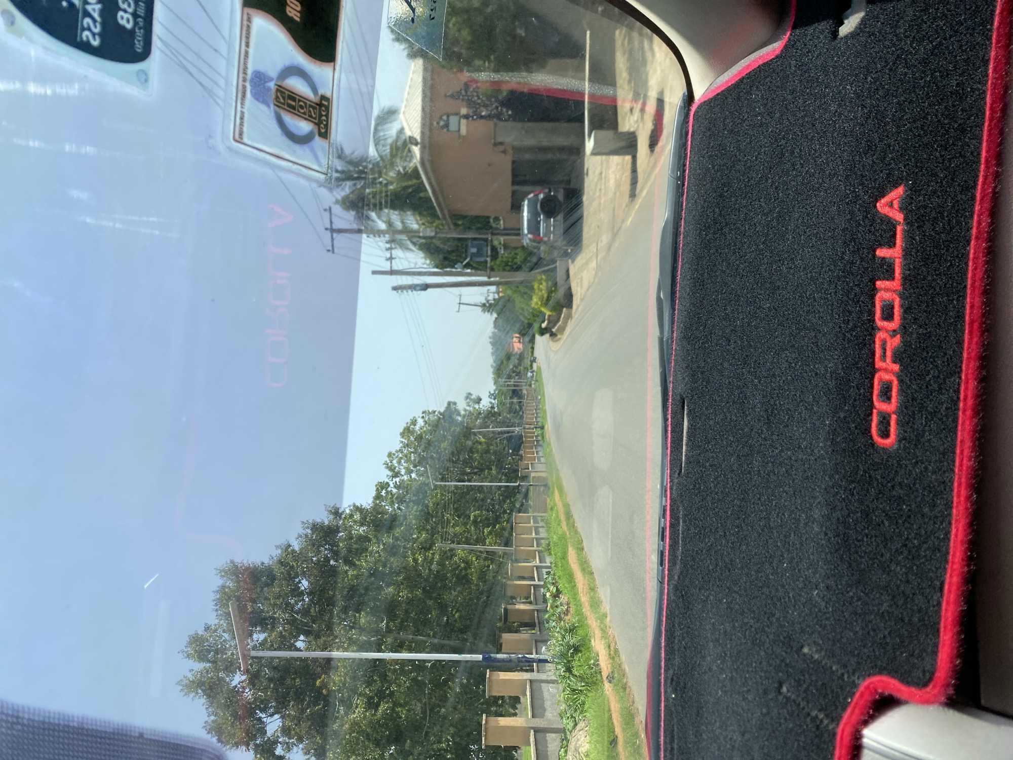 The image, taken from the interior of a vehicle, depicts a sunlit street scene in Bar Jirgi Summa, Nigeria. The foreground is dominated by a black dashboard mat with a red trim and the embossed red lettering "COROLLA". The windshield reveals a paved road lined with a low wall and a series of pillars on the left, followed by lush green trees and vegetation. On the right side of the road, a building with a brown facade is visible, with a dark-colored SUV parked in front of it. Utility poles with electrical wires are scattered along the street. The sky is a clear blue.