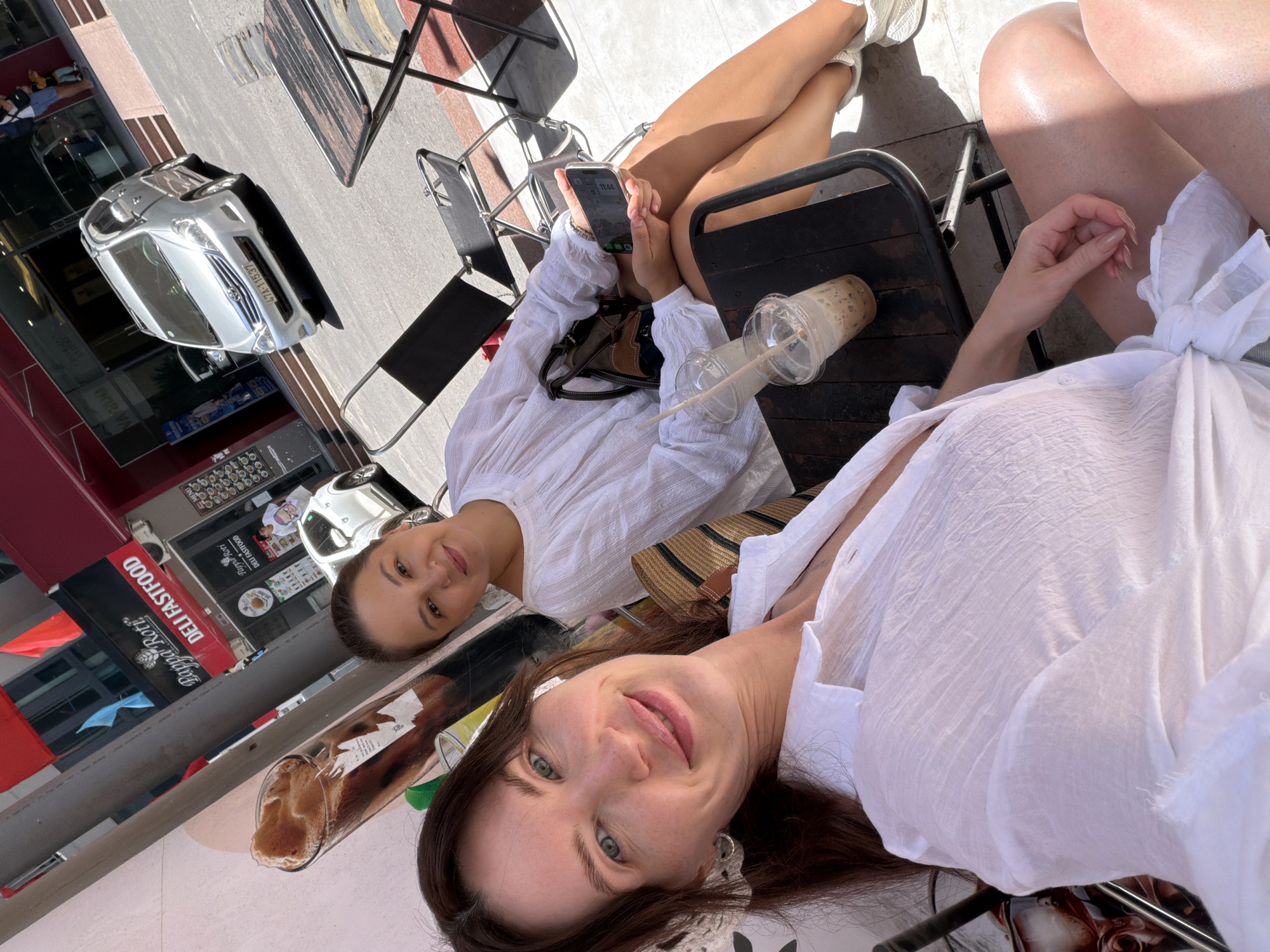 The image captures two women seated at an outdoor cafe or restaurant on a sunny day in what is identified as Bac Nha Trang Ward, Vietnam. The woman in the foreground, positioned to the lower left, has long brown hair and light eyes, and is taking a selfie. She wears a white, textured, collared shirt with long sleeves, which appears to be tied at the waist. White wireless earbuds are visible in her ears. Her fingernails are long and manicured with light polish.
Behind her, to the right, sits another woman with brown hair, smiling towards the camera. She wears a white, loose-fitting top with long sleeves. She holds a silver-colored smartphone displaying the time "11:43" in her hands and has a small, dark-colored handbag with tan accents resting on her lap. Her legs are visible, and she is wearing white sneakers. On the dark, rectangular outdoor table in front of the second woman are two clear plastic cups with straws; one contains an iced coffee with foam, and the other a lighter-colored beverage.
The background reveals a bustling street scene. Across the road, a silver Toyota Innova minivan with the Vietnamese license plate "47A-115.37" is visible. Behind the vehicle are commercial buildings featuring red awnings and glass storefronts. A prominent sign for "Pappa Roti DELI FASTFOOD" is clearly displayed on one of the buildings. Above the storefronts, red and blue banners or flags are mounted. A dark utility pole stands between the subjects and the background buildings. The scene is brightly illuminated by sunlight, creating distinct shadows on the ground and objects.