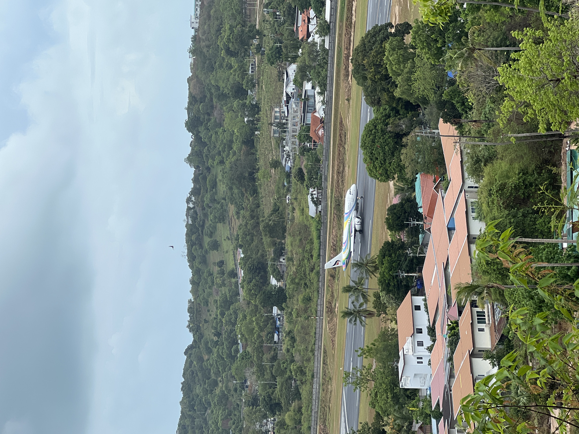 A vertical, high-angle shot shows a cityscape and an airplane on a runway, possibly at an airport. The location is suggested to be near บ้านบางรักษ์, Thailand. The sky is overcast. A forest-covered hill slopes down to a mix of residential houses and what appears to be an airport runway. The houses mostly have terracotta-colored roofs. The airplane is mid-run on the runway, and has a light fuselage with colorful tail livery. A dark bird flies in the sky near the hill. Green foliage frames the right of the shot.