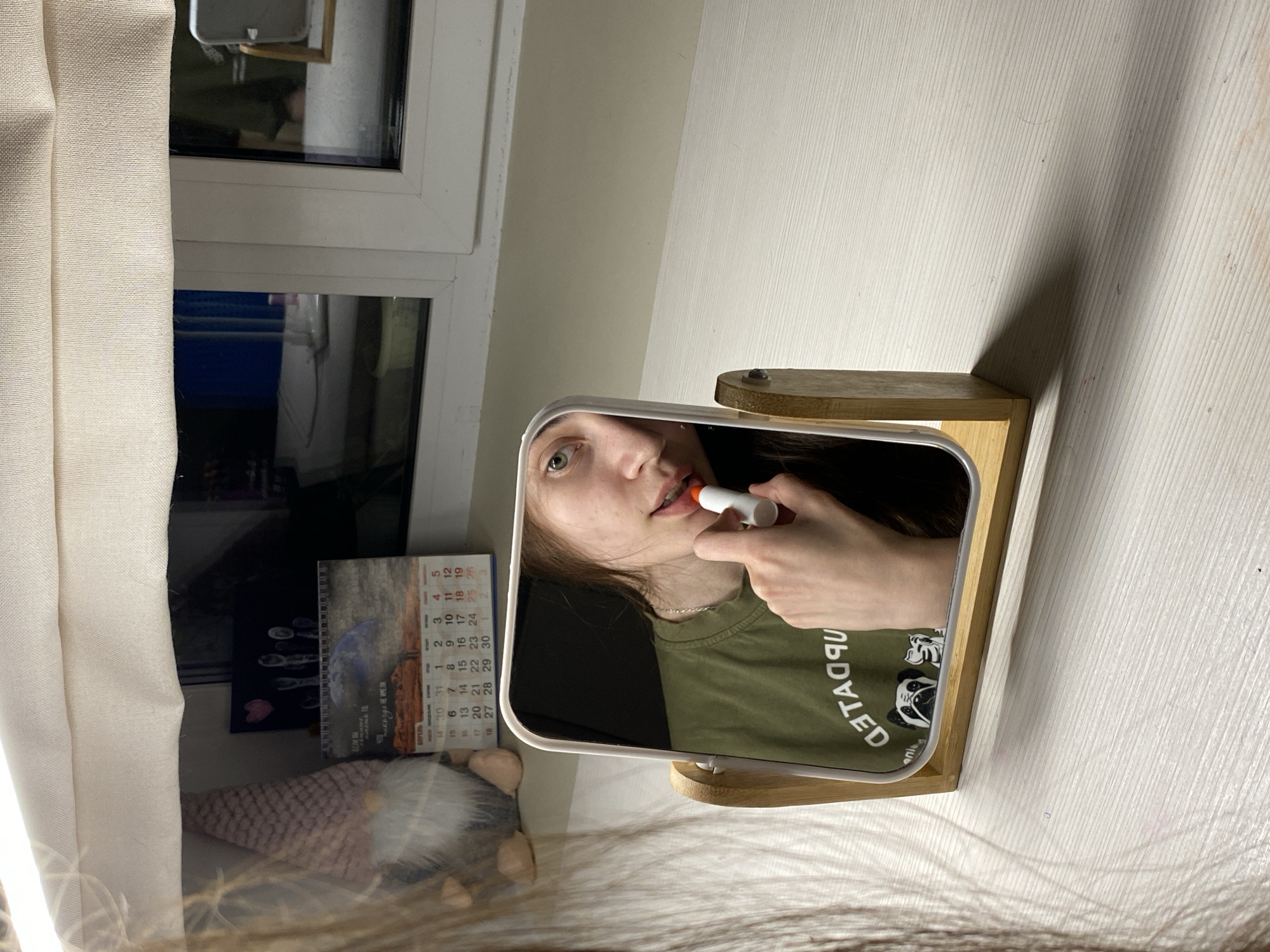 A person is seen in the reflection of a makeup mirror, applying lipstick. The mirror is placed on a wooden stand. To the left of the mirror is a window with a white frame, partially covered by a light-colored curtain. A calendar and a small gnome figurine are visible near the window. The wall behind the mirror is off-white. Based on the calendar text, the scene is likely located in the city of Votkinsk, Russia.