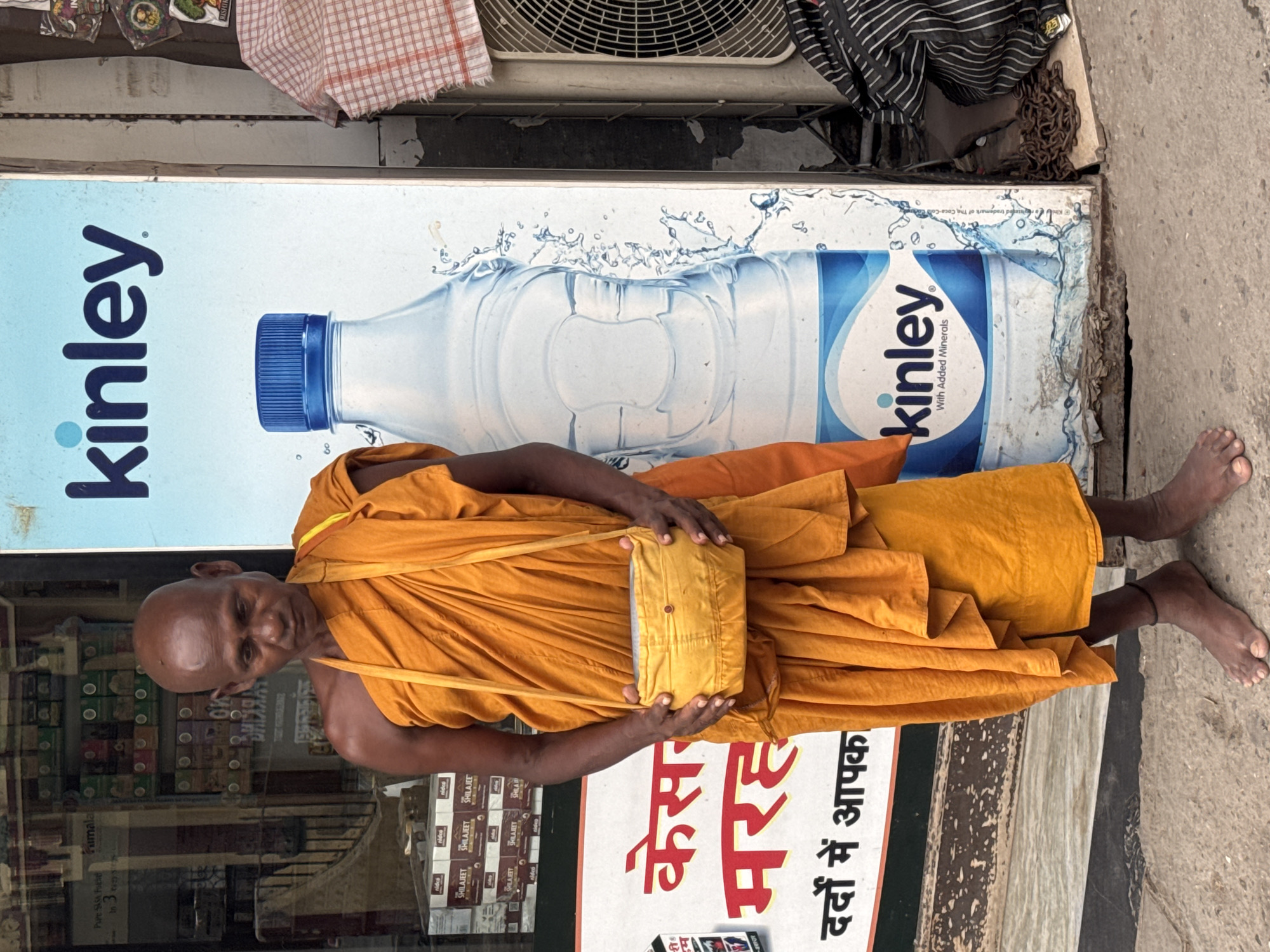 A bald, middle-aged man wearing an orange robe stands barefoot on a concrete sidewalk in Rishikesh, India. He has a yellow pouch slung across his chest. Behind him is a large advertisement for Kinley bottled water, featuring a picture of a water bottle with a blue cap. To the left of the man is a storefront displaying various products, including boxes of what appears to be herbal or medicinal items. To the right of the advertisement, a piece of checkered cloth is draped over an air conditioning unit, and a metal chain lies on the ground. The scene appears to be outdoors in a commercial area of the city.