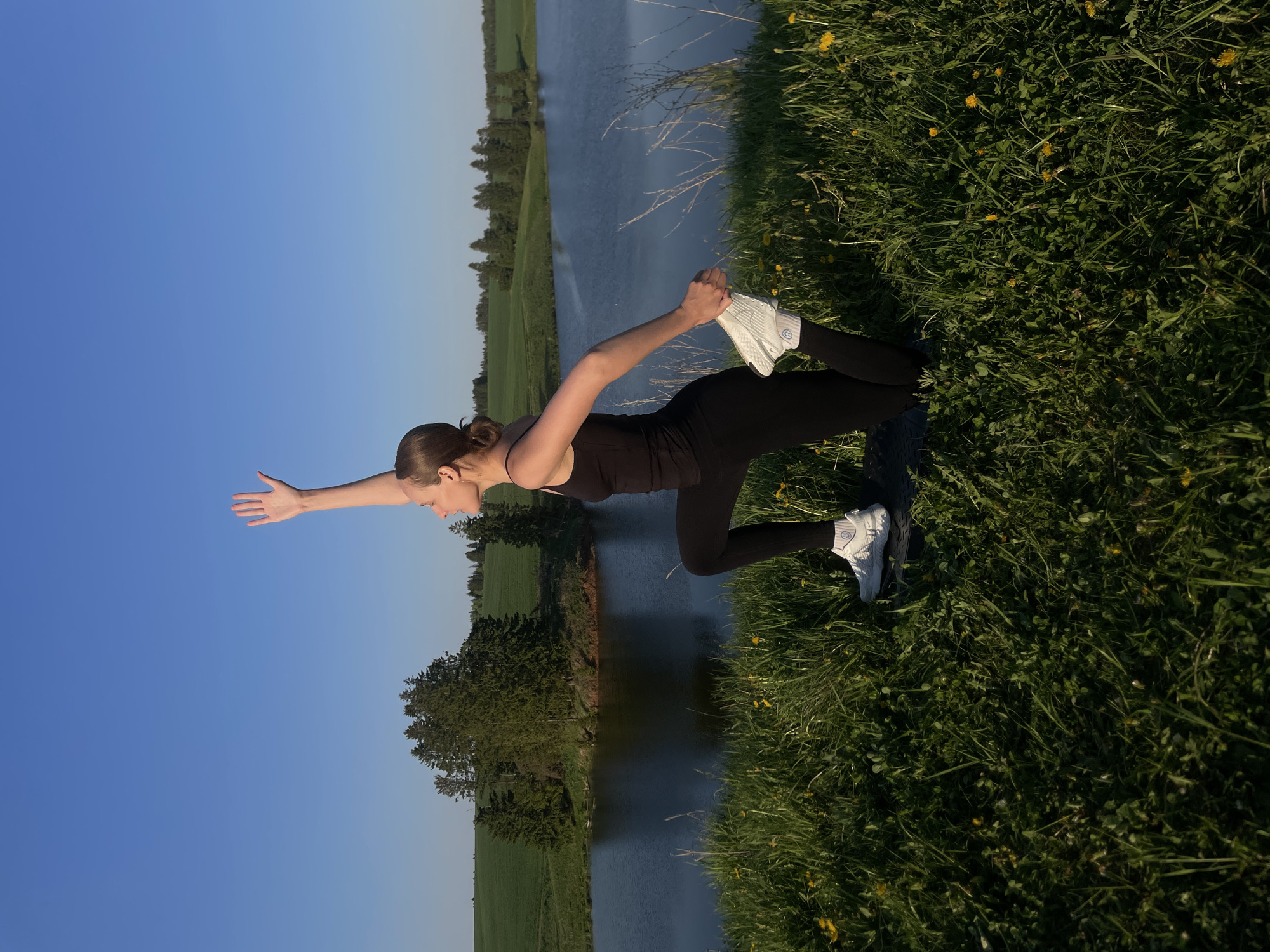 A woman is stretching outdoors. She is kneeling with her left knee on the ground and her right foot in her right hand, extending her left arm. She is wearing a black tank top, black leggings, and white sneakers. She is positioned near a body of water, with green fields and trees on the opposite bank. The sky is clear and blue. The ground around her is covered in green grass and yellow flowers. The location is outdoors. There is no information about city name.