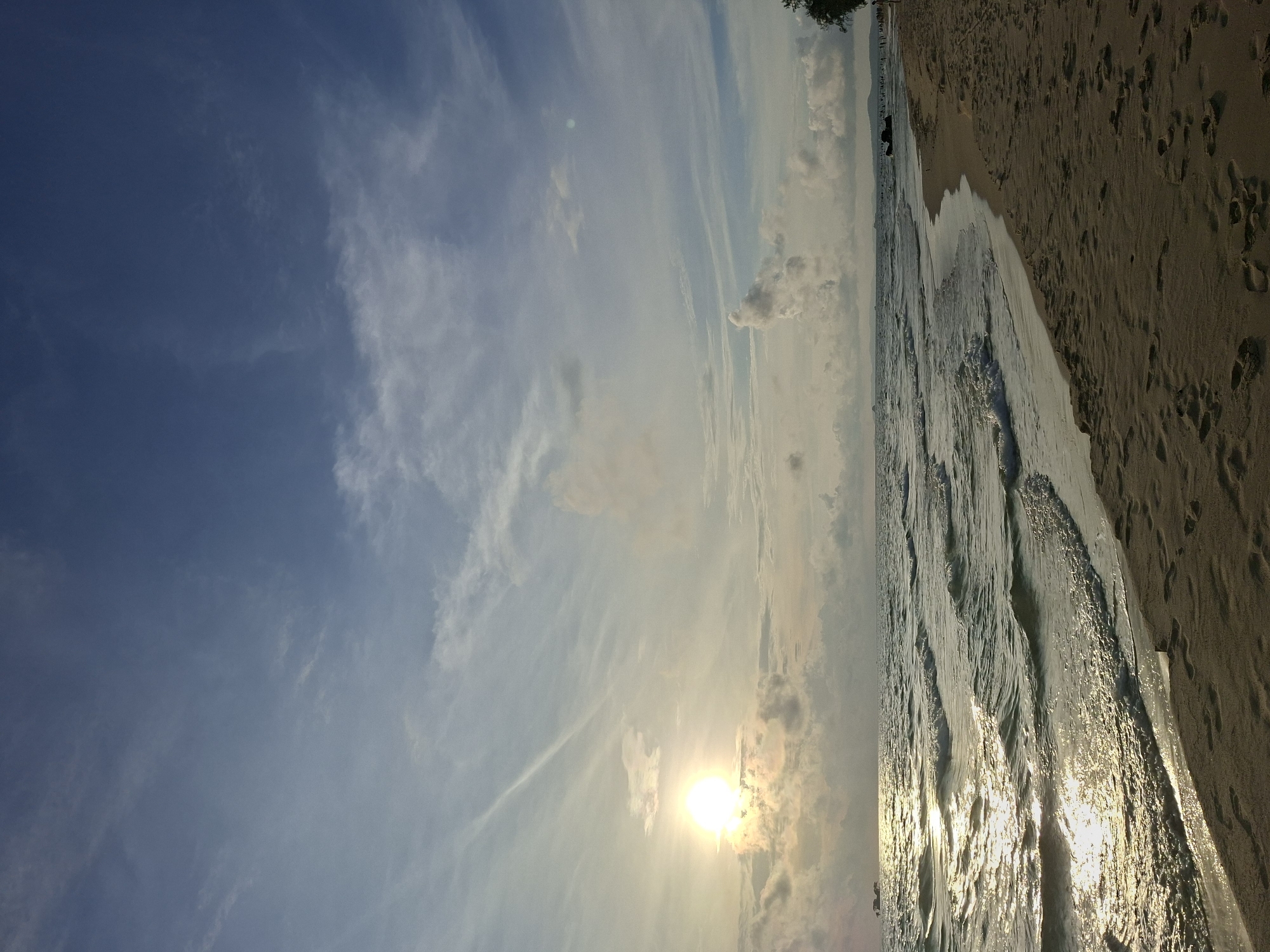 A wide shot of a beach at sunset shows the sun low on the horizon, casting a golden sheen on the rippling ocean waves. The sky is a gradient of blue, with wispy white clouds scattered across it. The foreground is a sandy beach with footprints visible. The ocean meets the shore with gentle waves. In the distance, a few indistinct figures can be seen on the beach and in the water, along with a dark object in the water that may be a boat. The scene appears serene and natural.